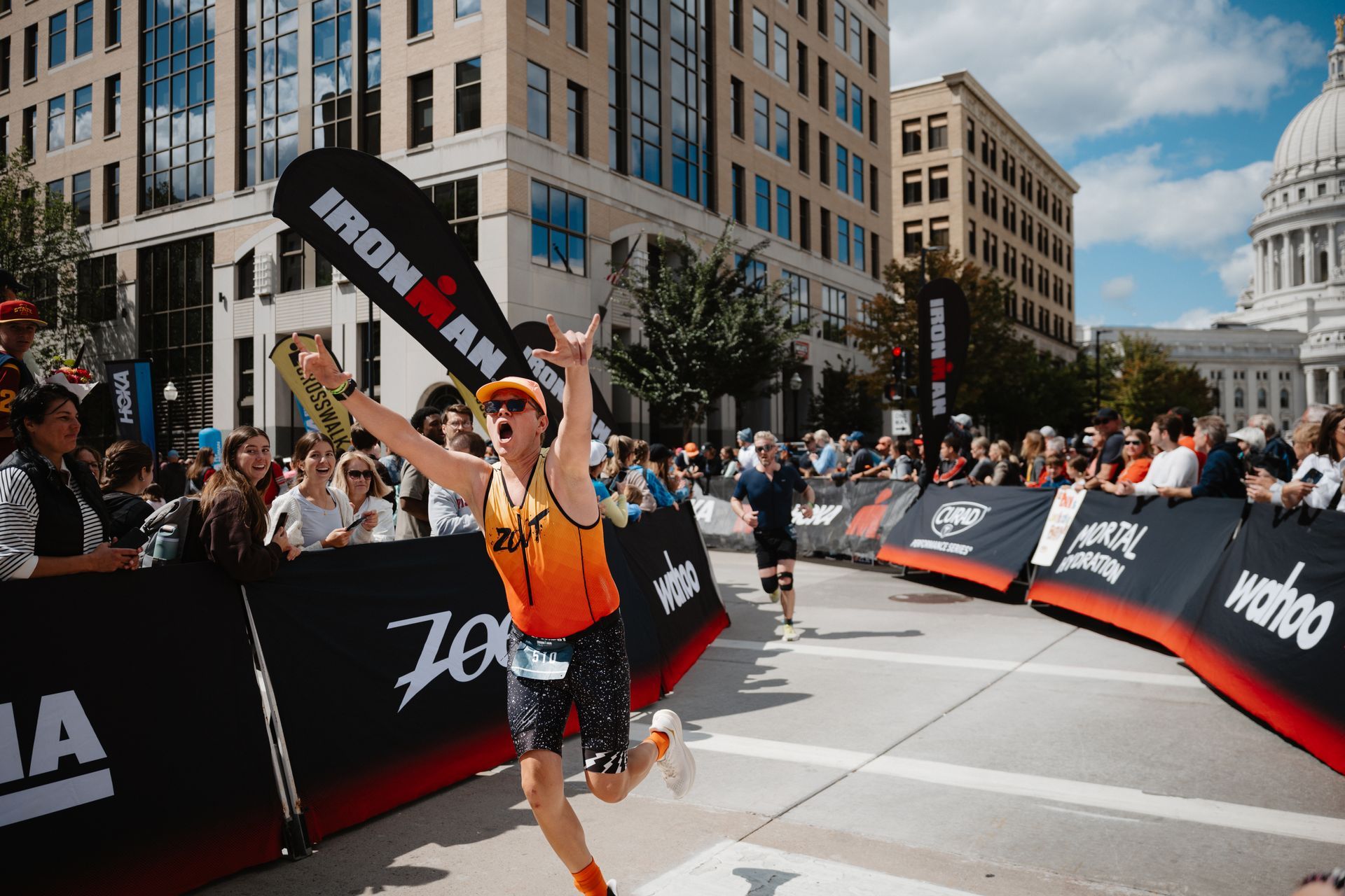 Runner holding a baton crosses the finish line at a city marathon, with spectators and the U.S. Capitol in the background.