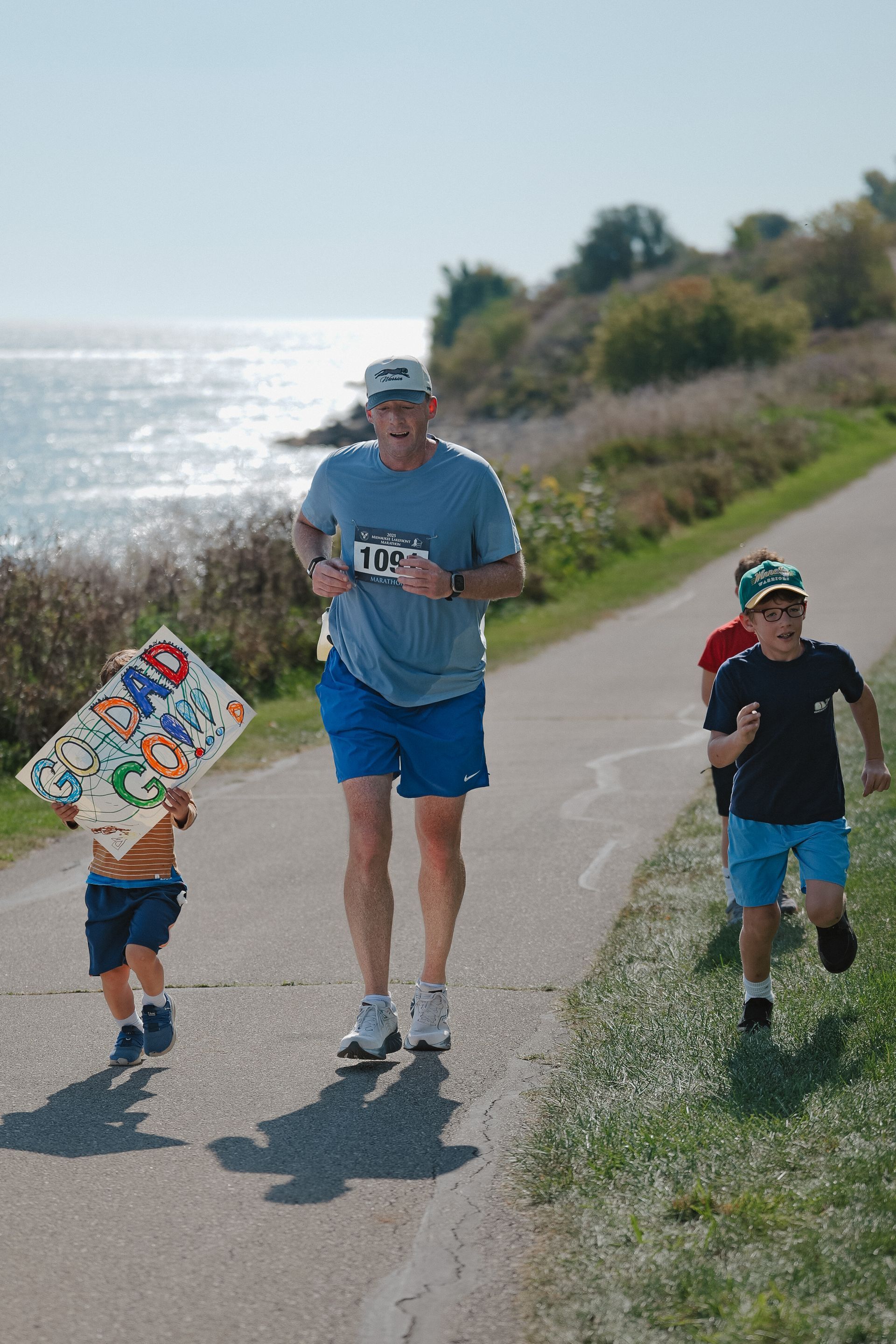 Three people jogging on a coastal path by the water, with one child carrying a colorful sign.