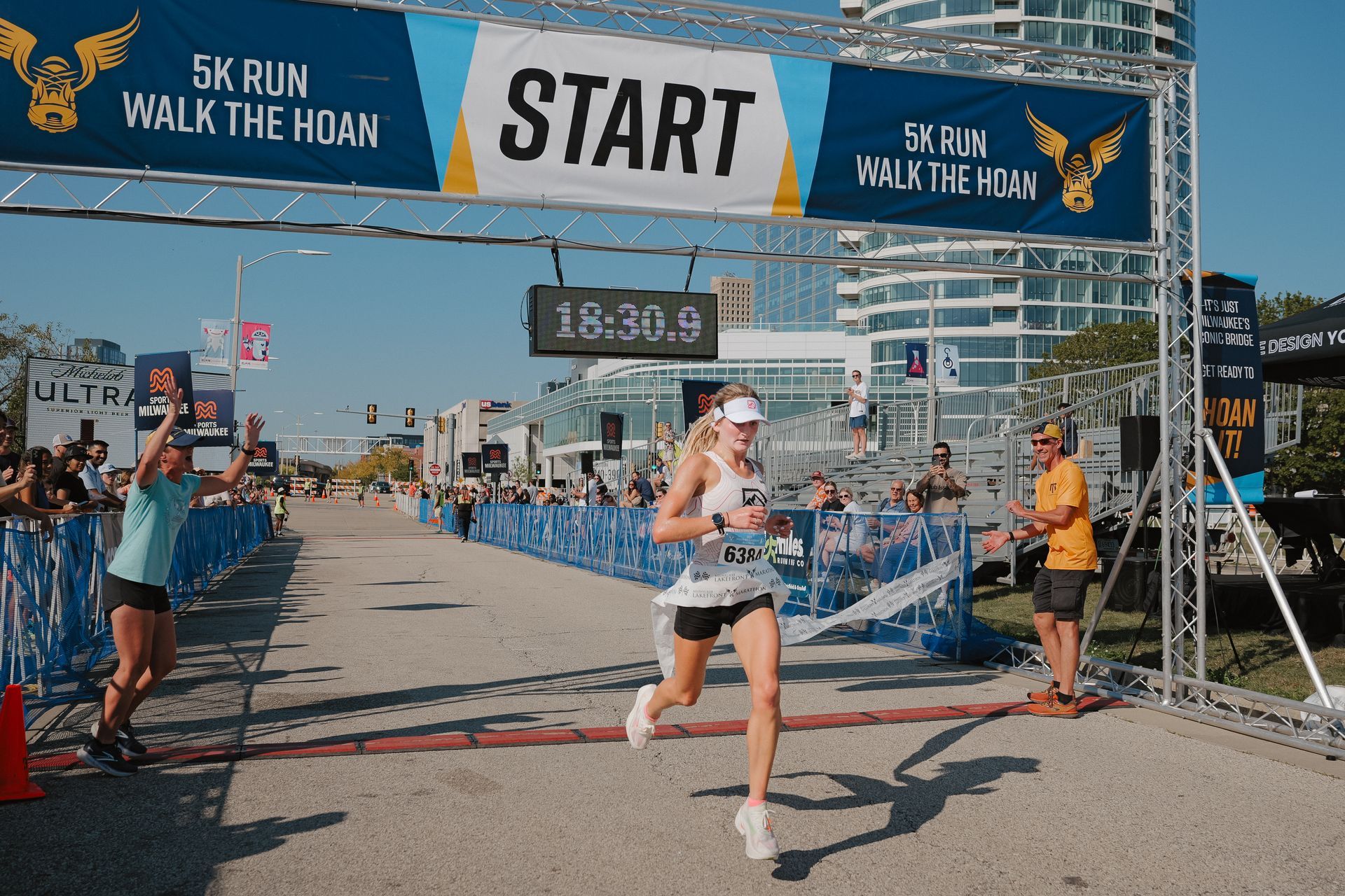 Runner crossing start line at 5K run/walk event under blue START banner, with spectators and city backdrop