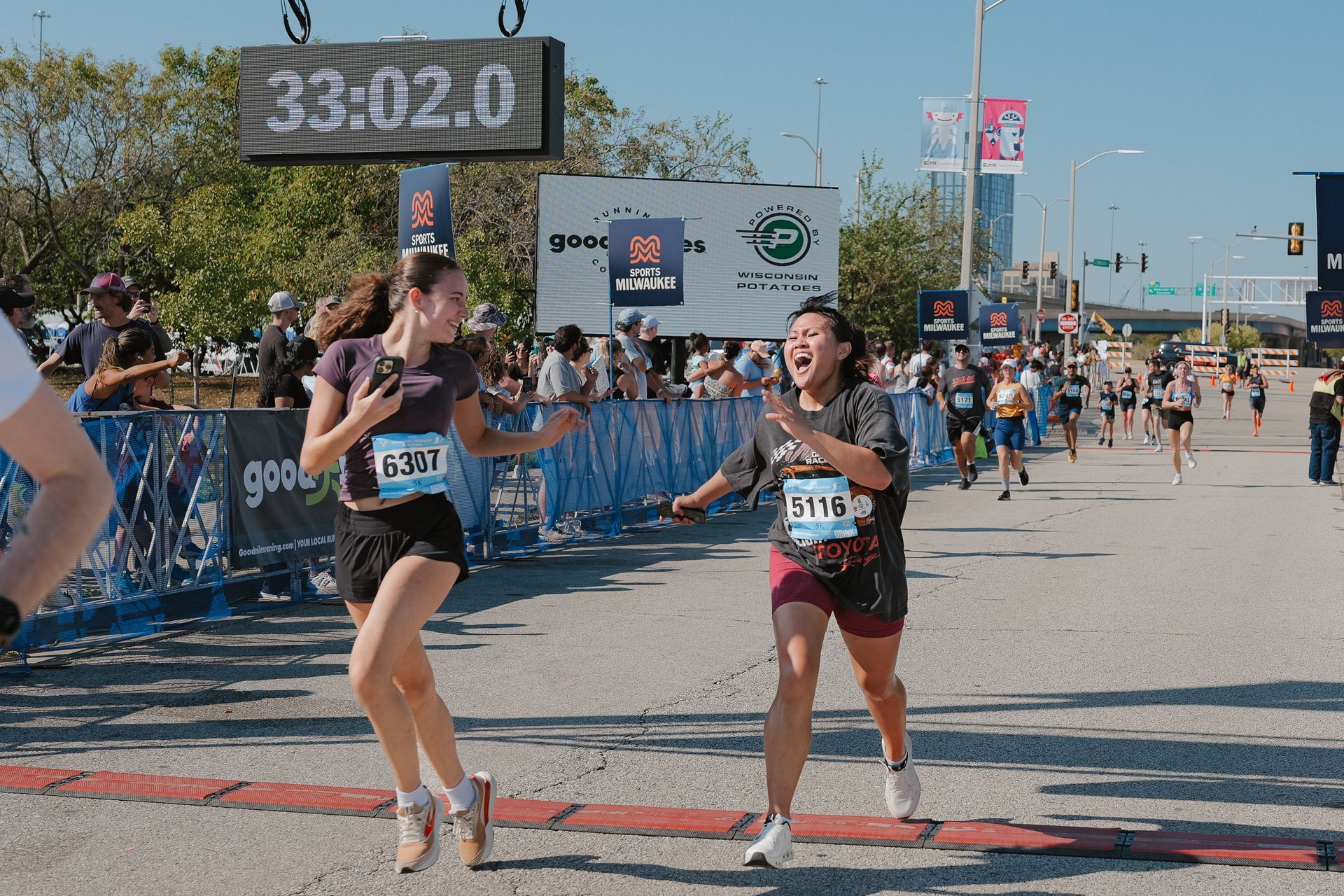 Runners sprint toward a finish line at a road race, with a timer overhead and spectators behind barriers.
