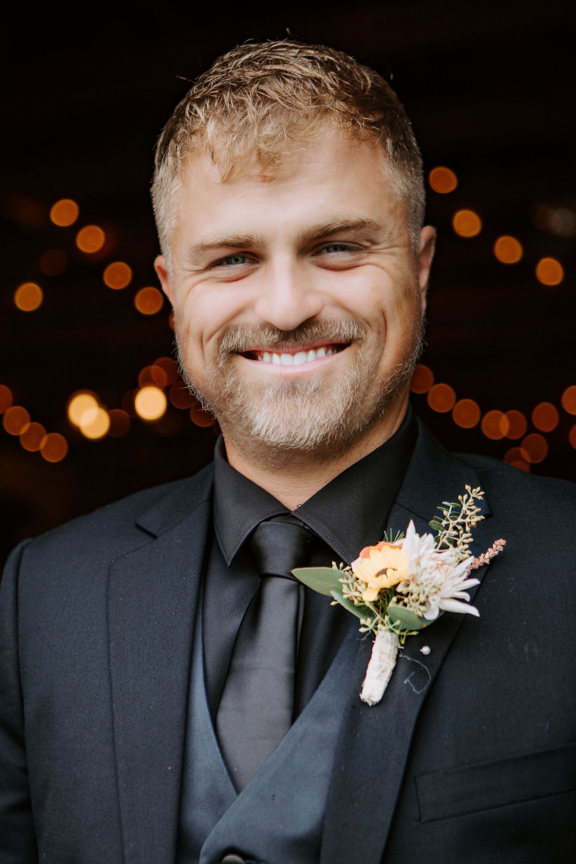 Smiling man in a black suit and tie with a floral boutonniere, standing against warm blurred lights