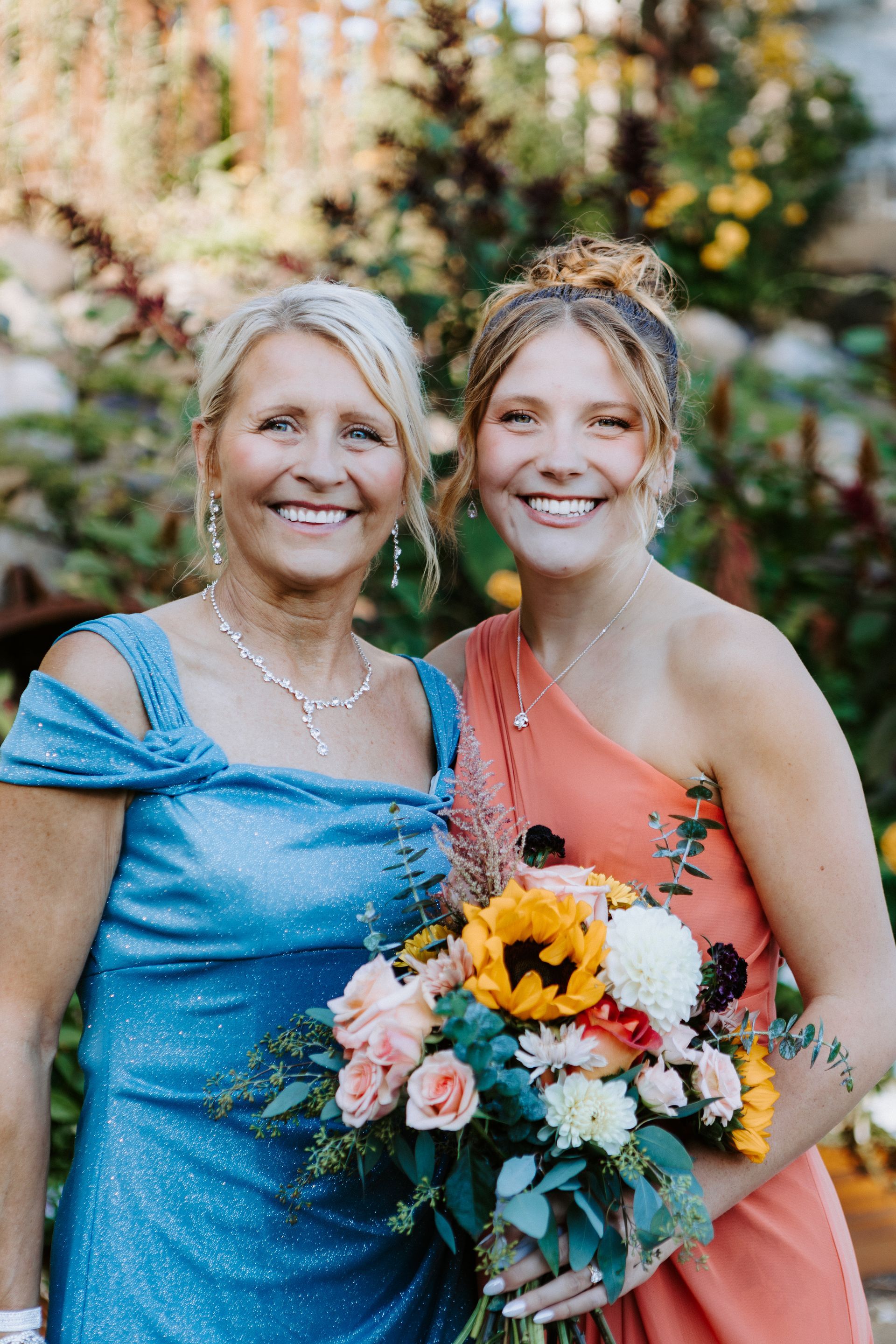 Two smiling women in formal dresses, one holding a colorful bouquet, standing outdoors.