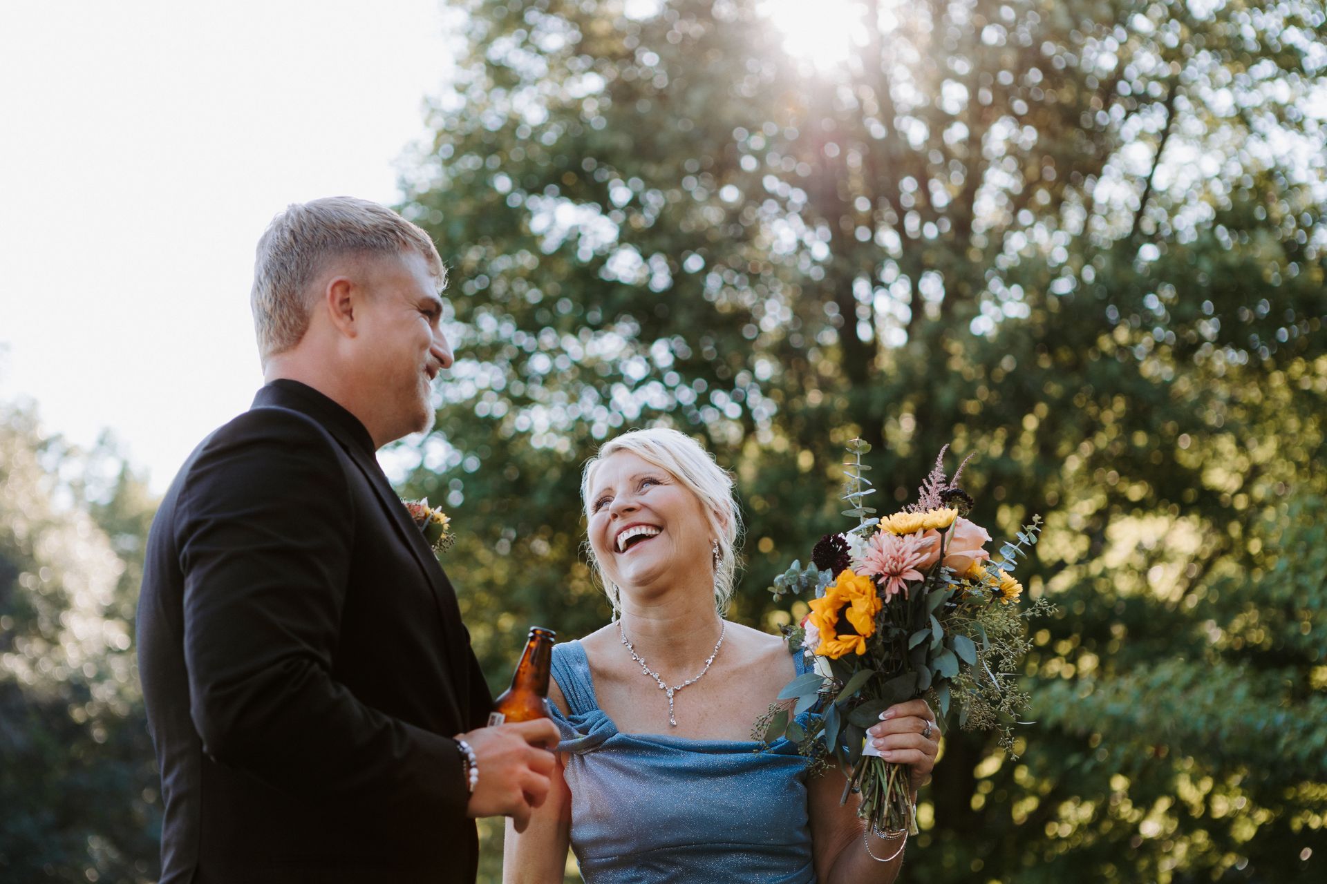 Two people smiling outdoors, one holding a bouquet of flowers at sunset.