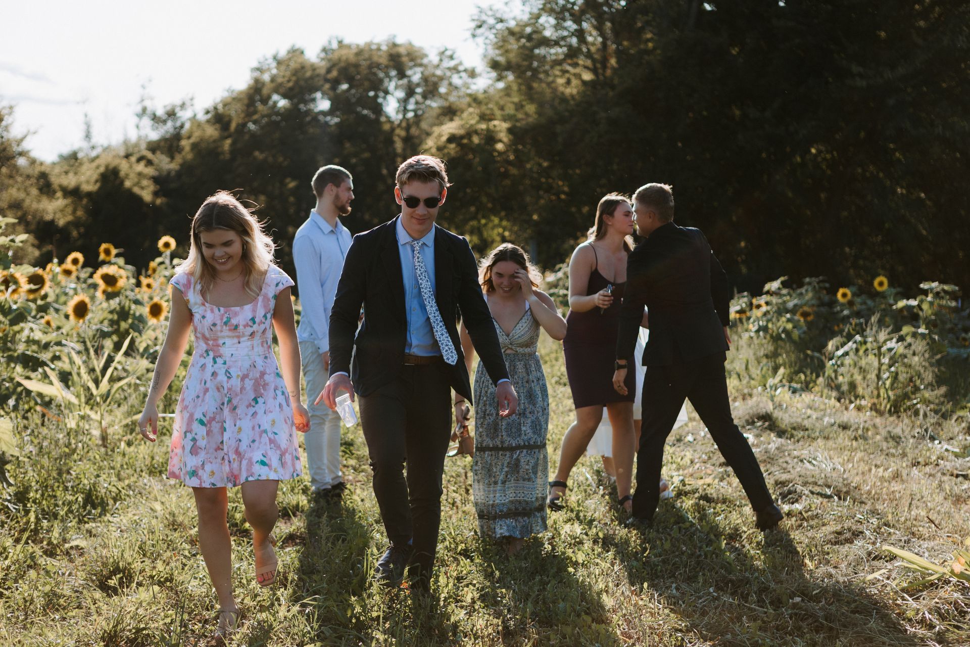 Five people walking through a sunny wildflower field, with sunflowers and trees in the background.