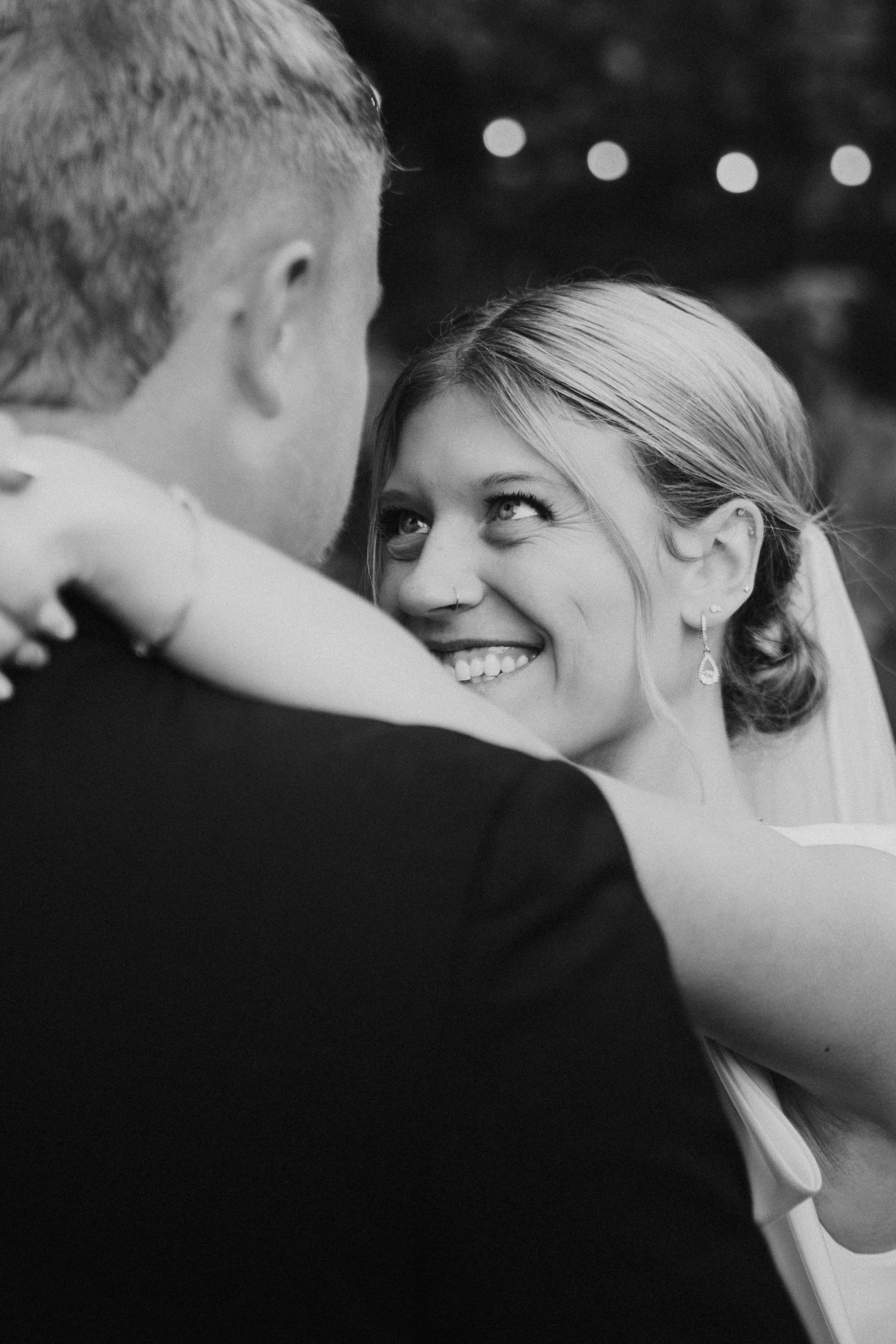 A smiling couple dances closely in a black-and-white photo with soft lights in the background.