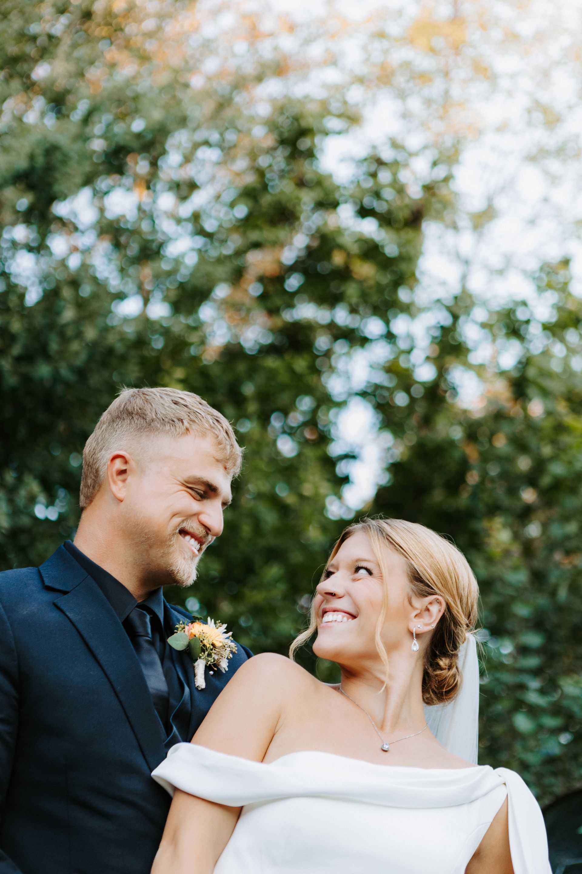 Smiling couple in wedding attire outdoors, looking at each other against a blurred green background