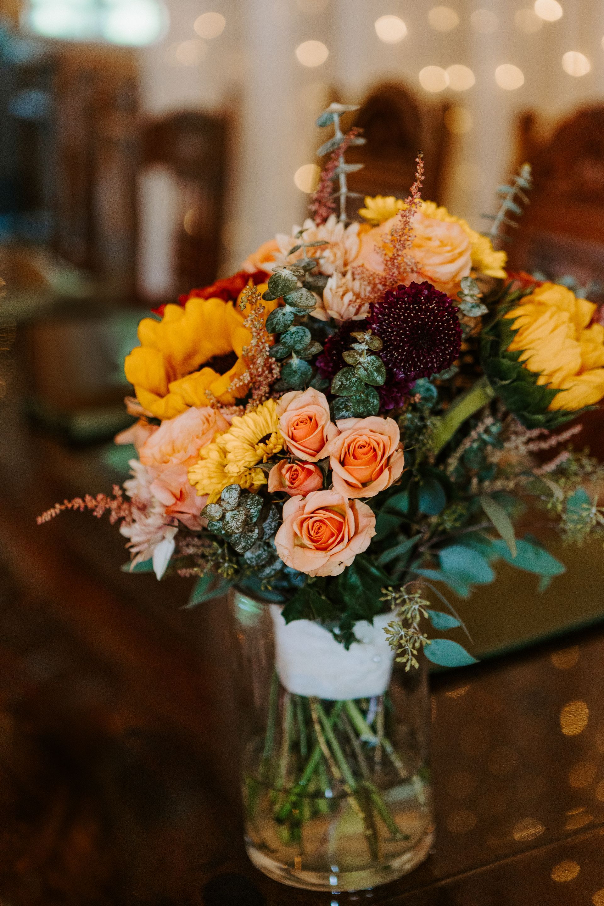 Rustic bouquet of yellow, peach, and burgundy flowers in a glass vase on a table
