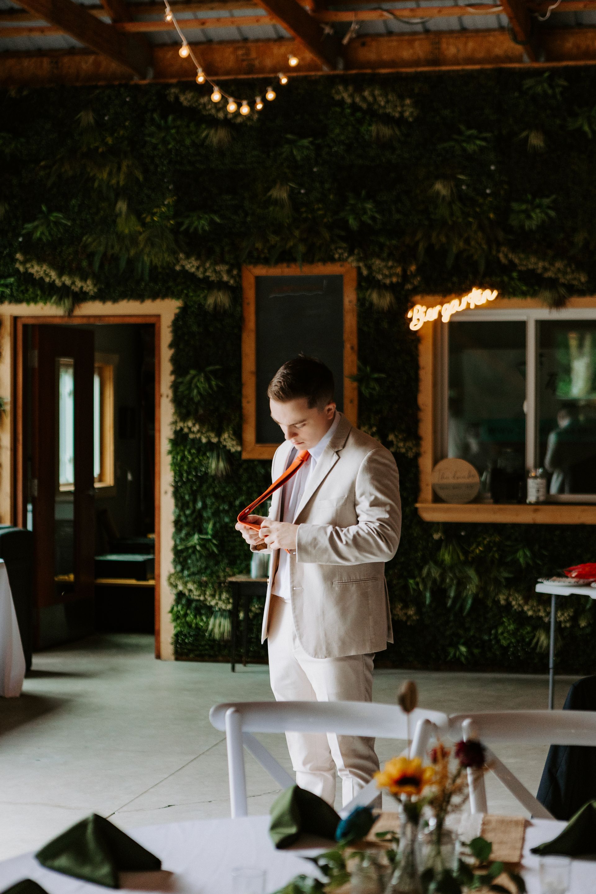 Person in a light suit standing in a rustic indoor venue, looking at a phone near a table with flowers