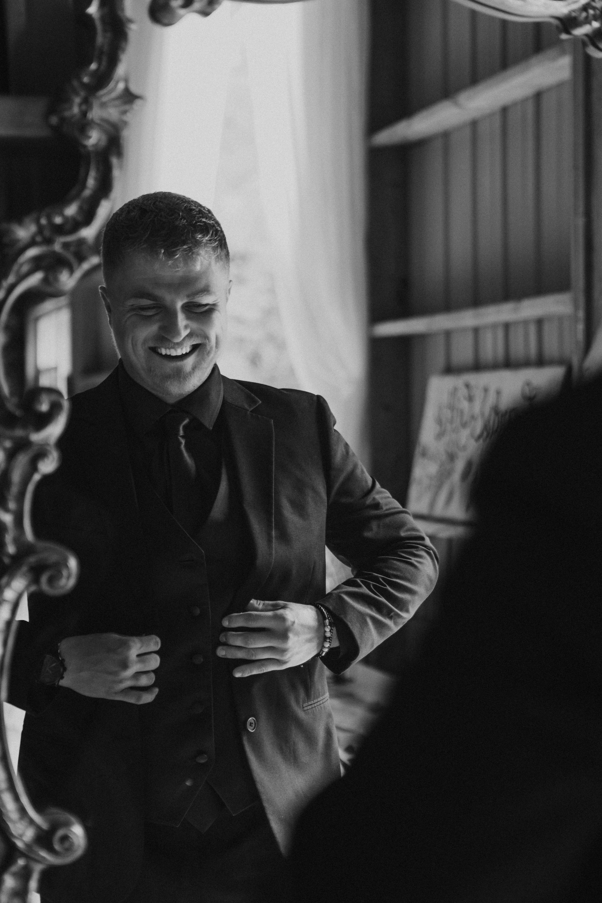 Man smiling while adjusting his jacket in a dimly lit room, seen beside a mirror.