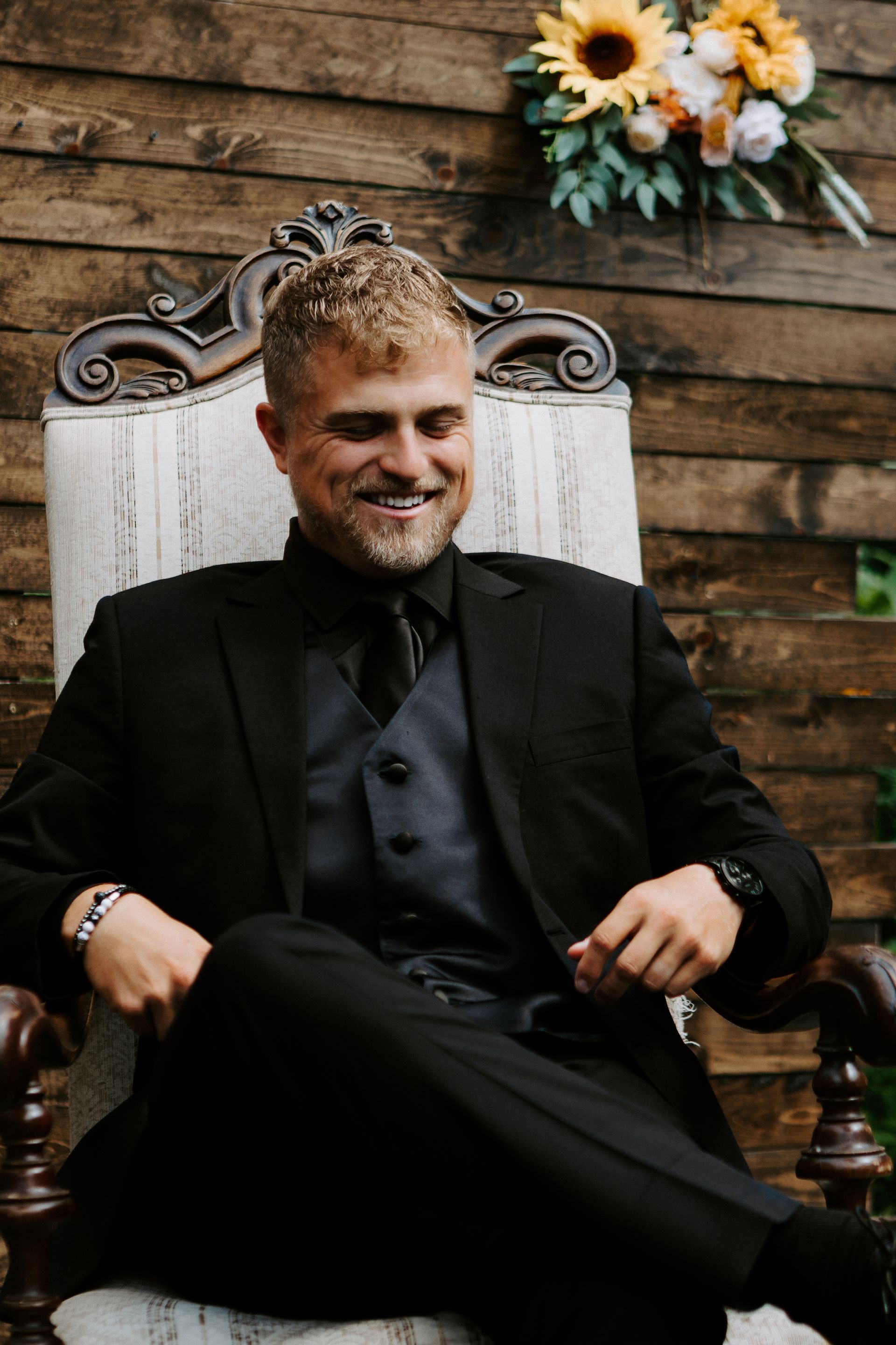 Man in a black suit sitting on an ornate chair, smiling against a rustic wooden backdrop with flowers.