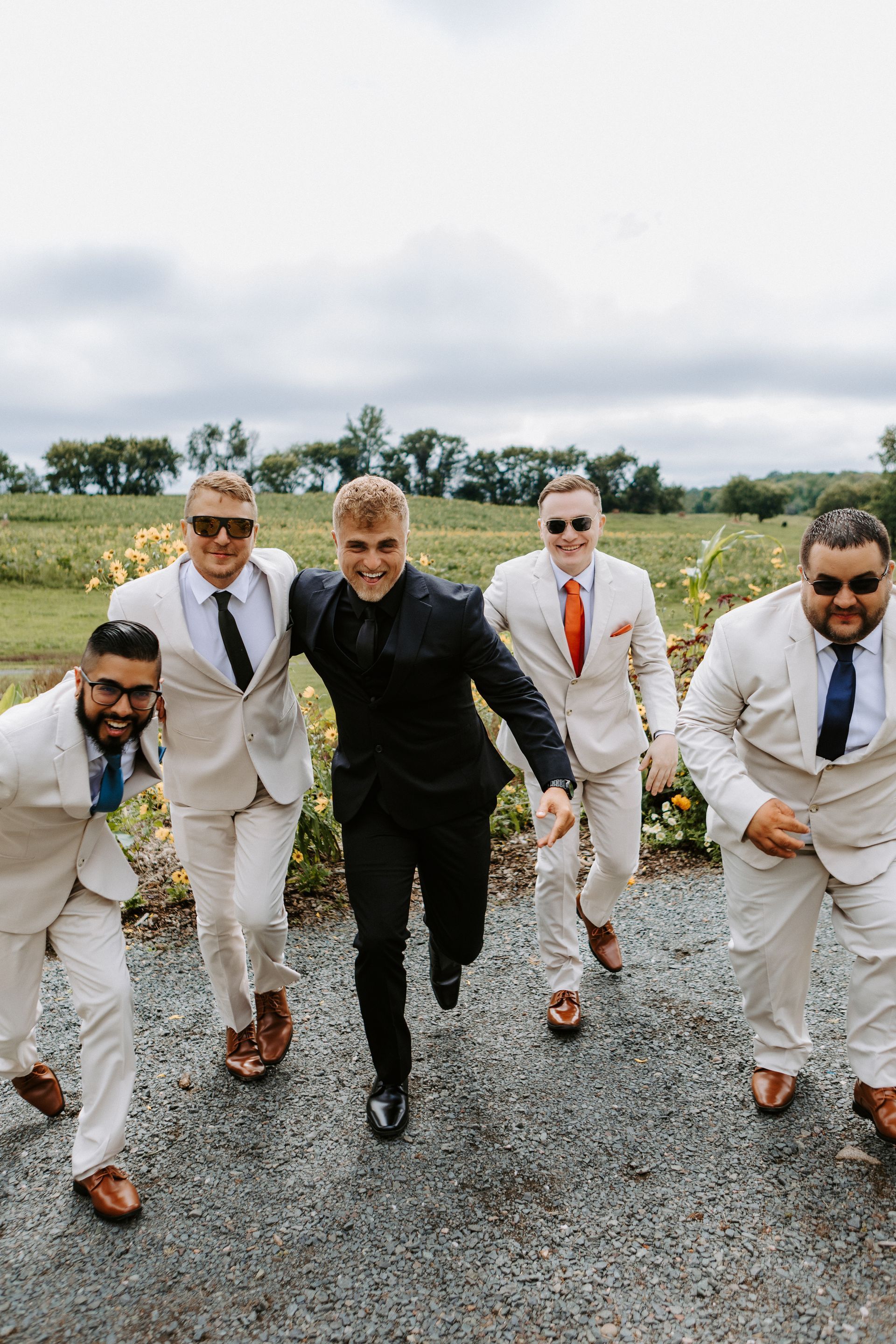 Five men in suits running toward the camera on a gravel path outdoors under a cloudy sky
