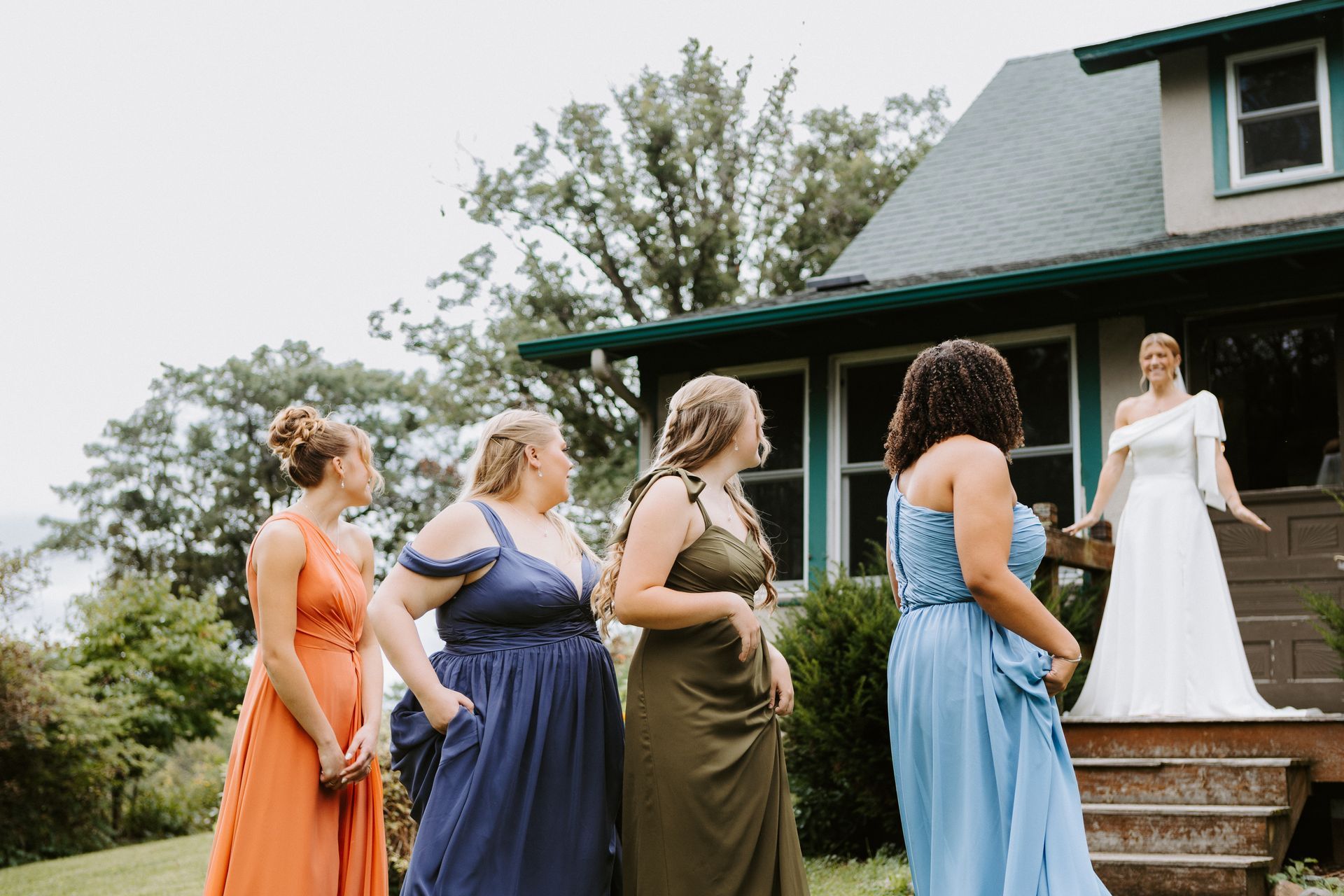 Wedding guests facing a bride on porch steps outside a house, with bridesmaids in colorful dresses in foreground
