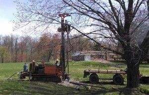 Orange drilling rig in a grassy yard beside a tree and utility trailer under leafless trees