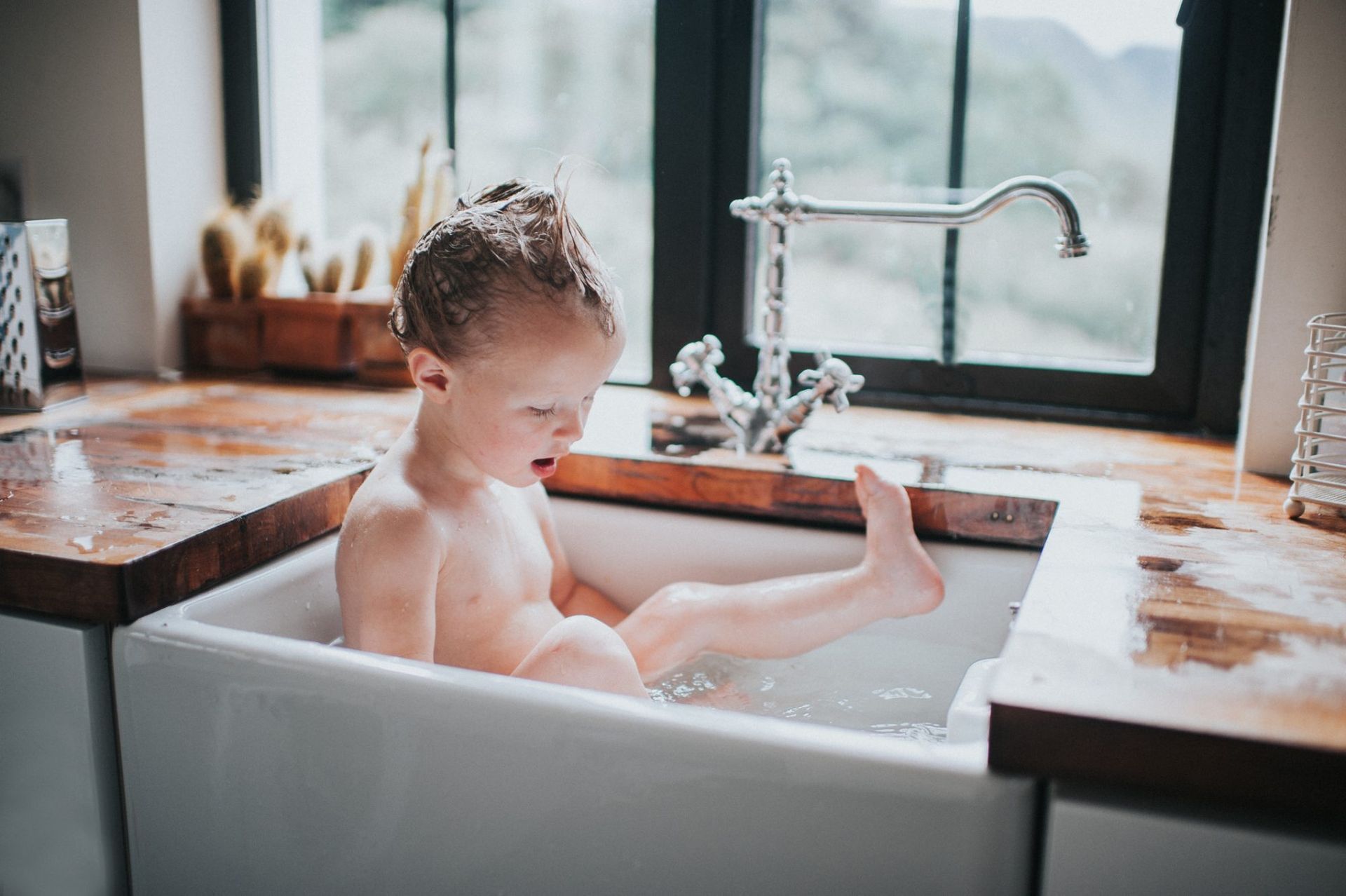 Child in a white bathtub by a window, splashing water in a bright kitchen