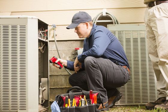 Technician crouches beside an outdoor HVAC unit, using a red tool near exposed wiring.