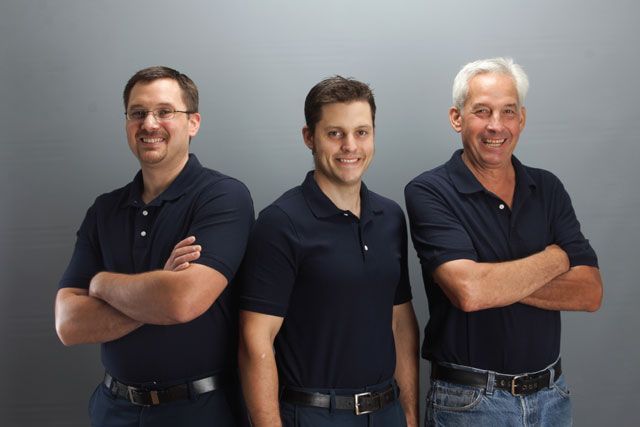 Three men in dark polo shirts and jeans standing with arms crossed against a gray background.