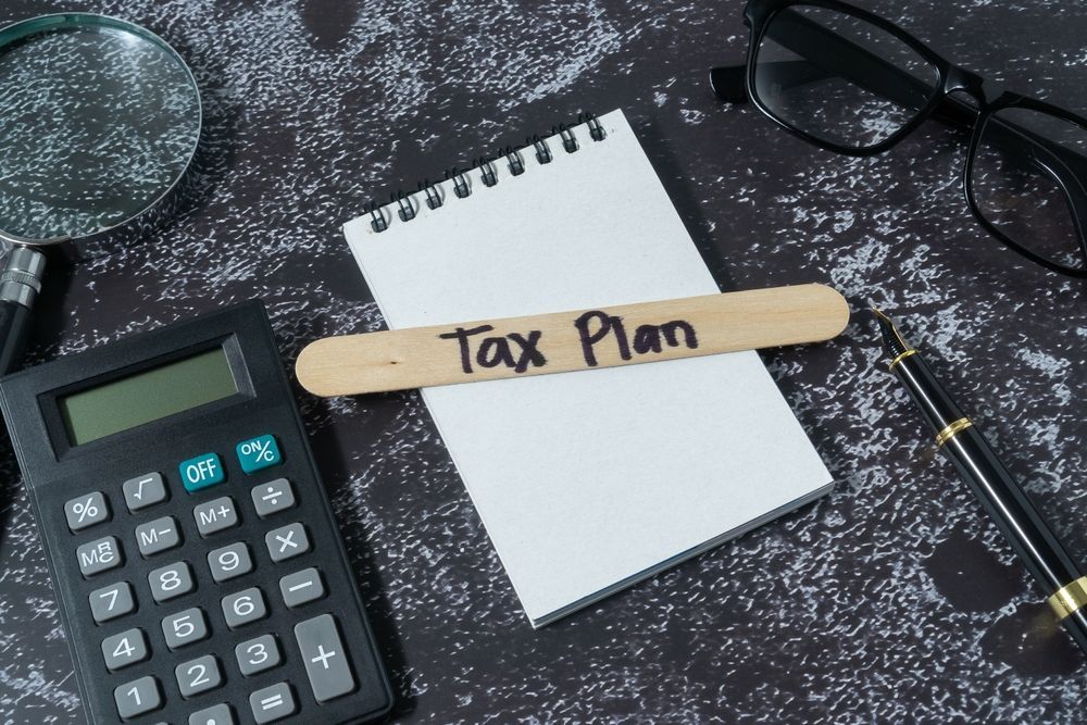 Calculator and notebook with “Tax Plan” label on dark desk, with glasses and pencil nearby