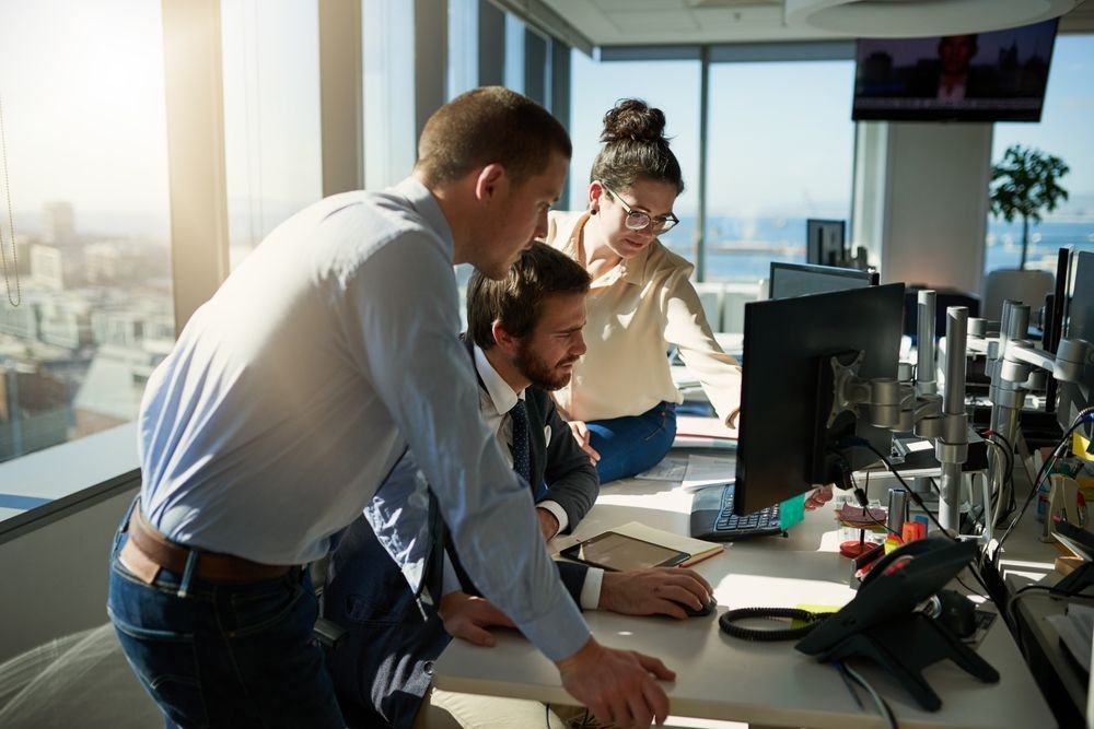 Coworkers gathered around a desk in a bright office, reviewing a computer monitor together.