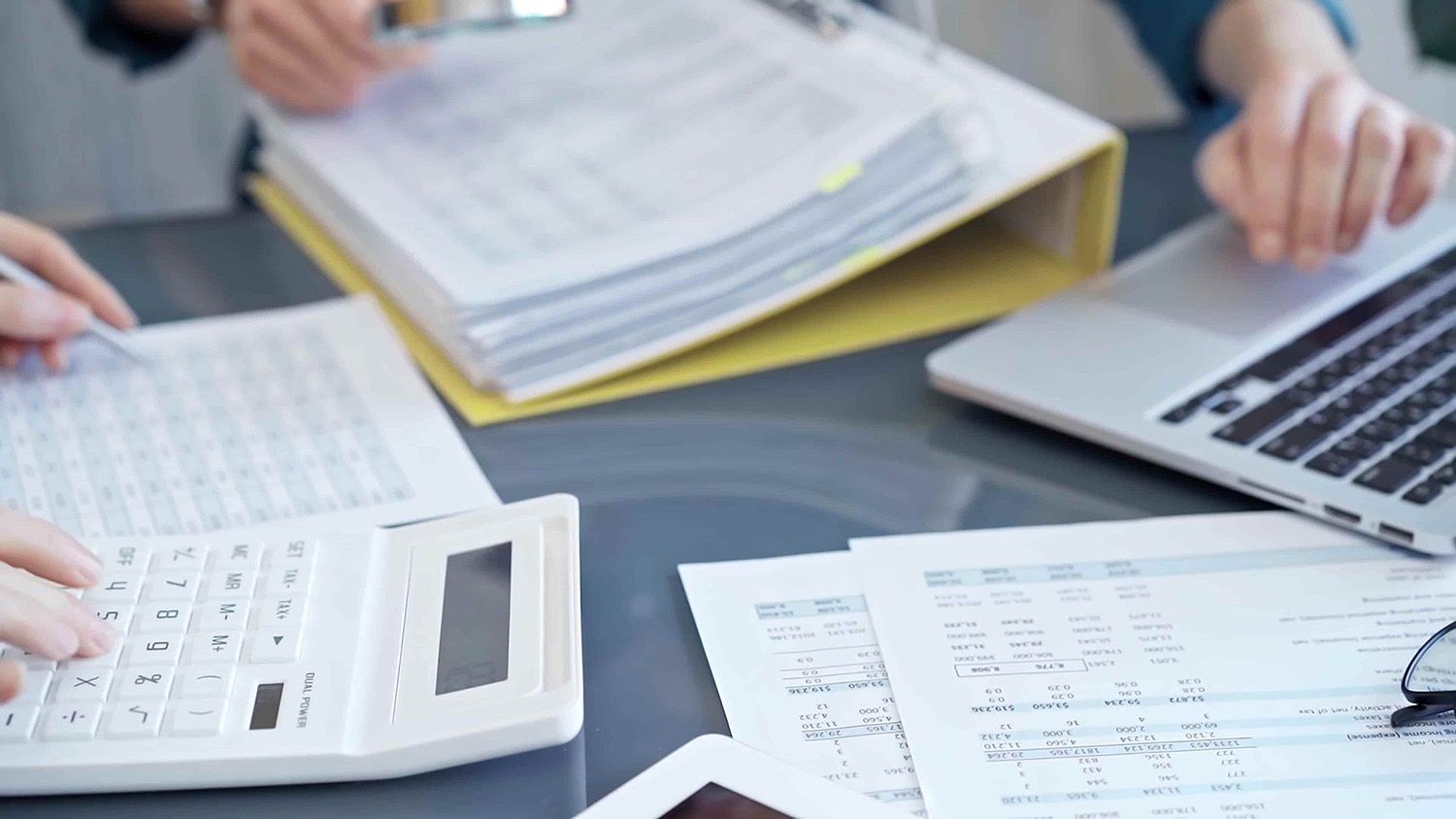 Hands Working on Accounting Documents with a Calculator, Laptop, and Binder