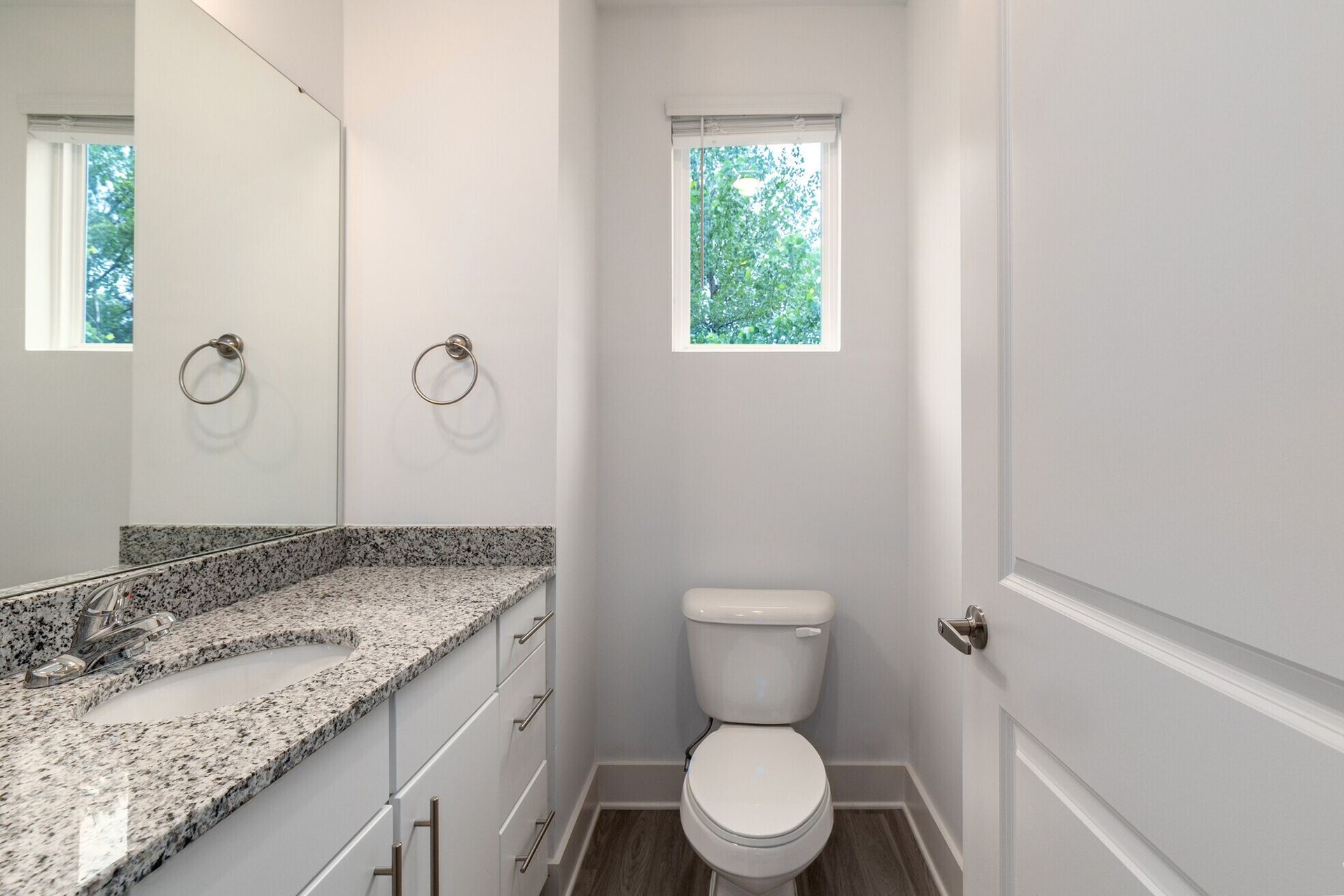 Small, white bathroom with a toilet, sink, and window; gray countertop, neutral walls, and dark flooring.