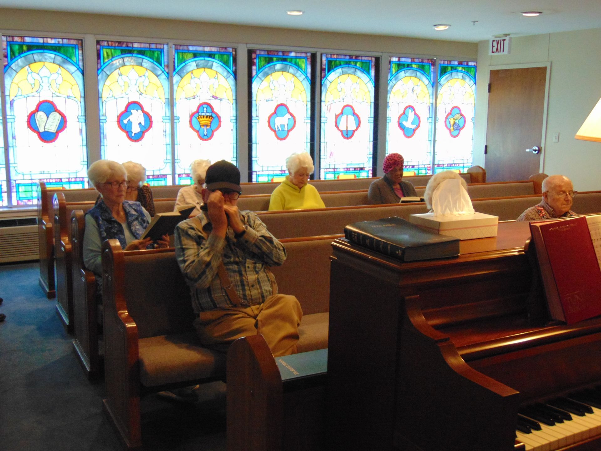 People seated in a church pew beside a piano, with stained-glass windows in the background.