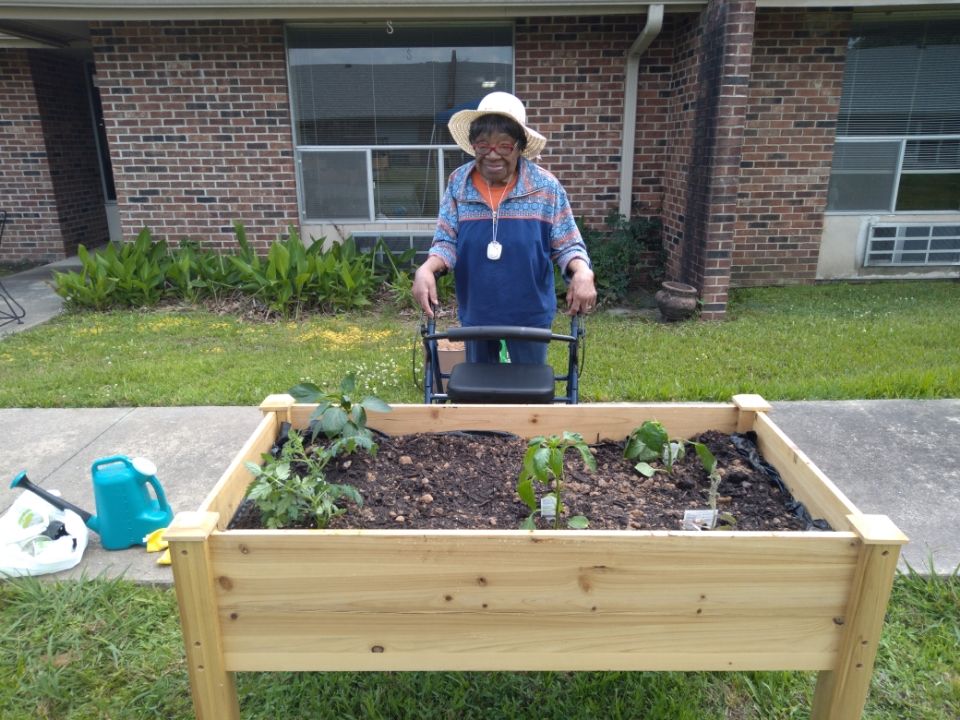 Person gardening beside a raised garden bed in front of a brick building, holding a watering can.