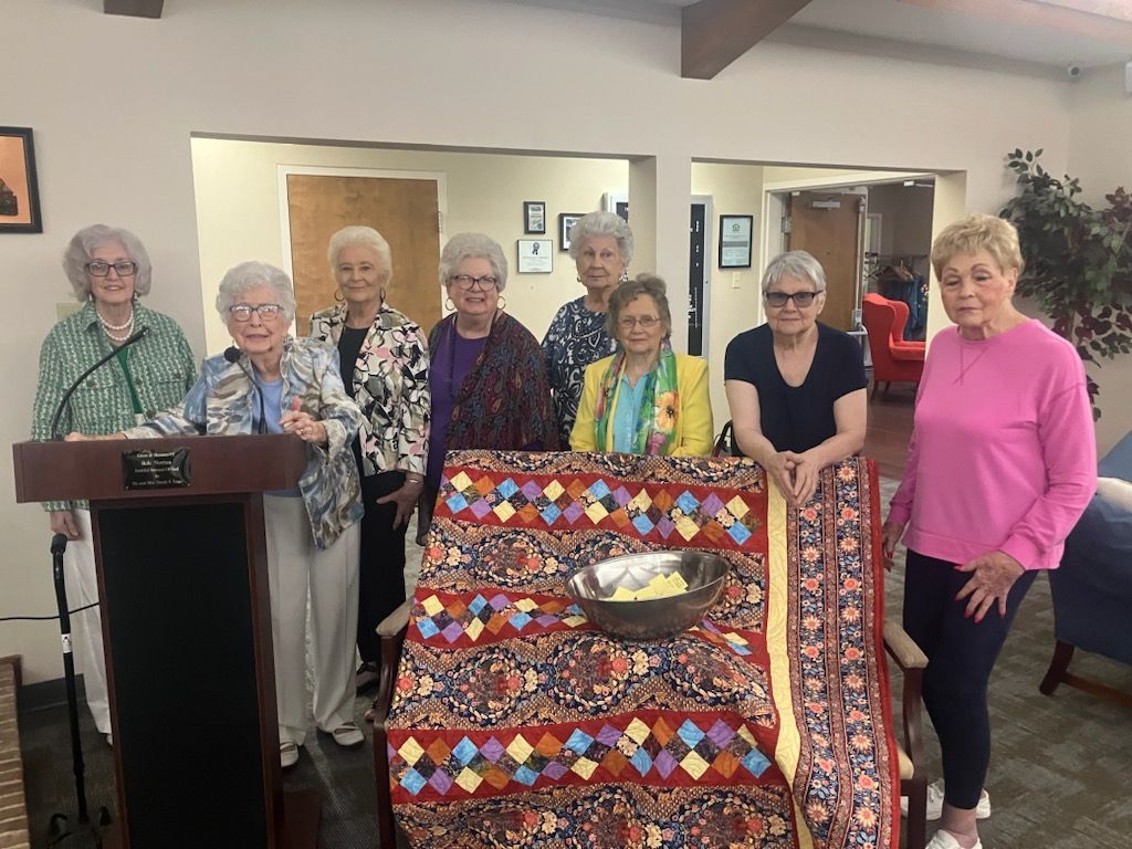 Six people stand indoors behind a colorful quilt display, with a podium at left.