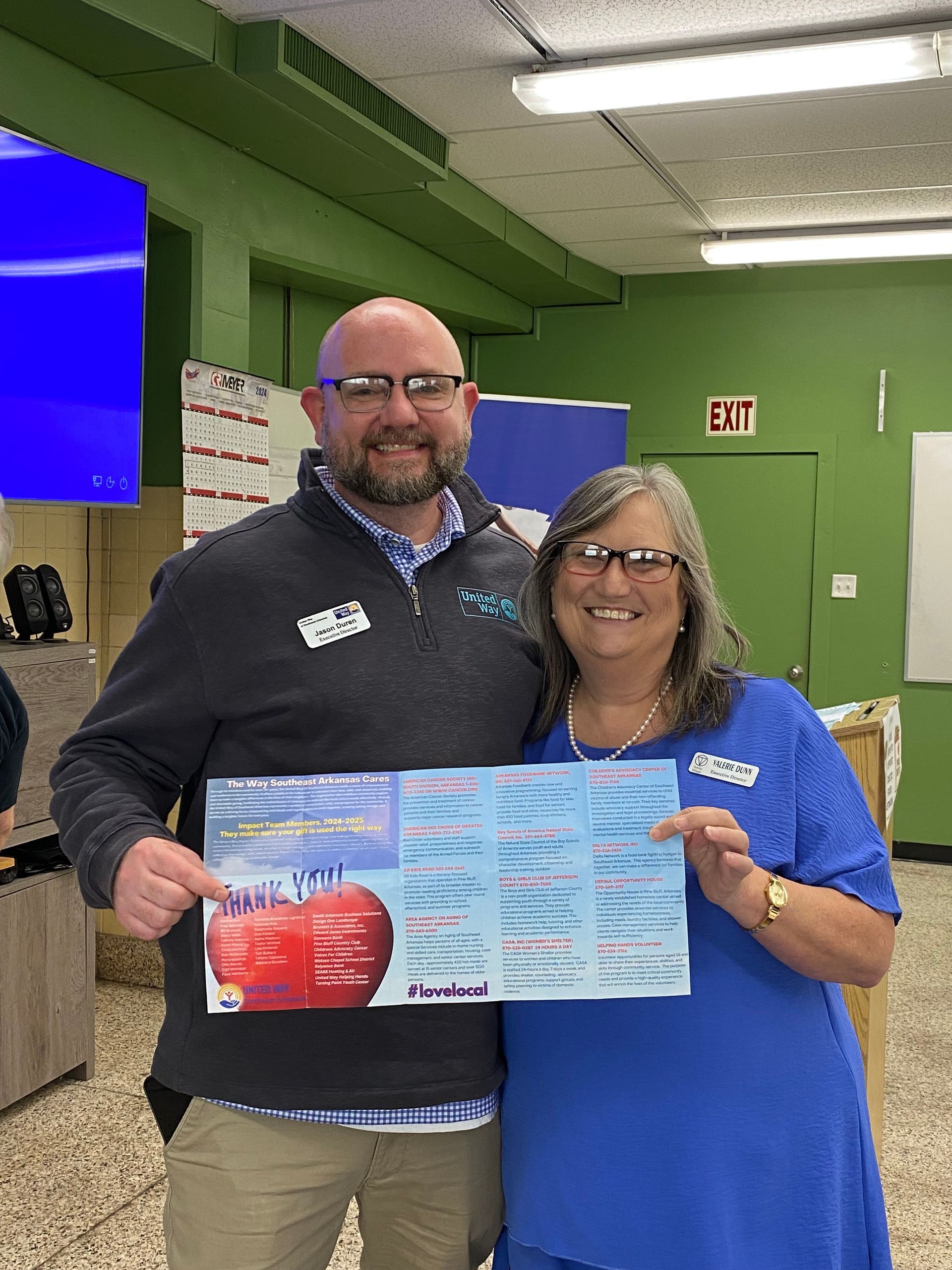 Two people holding a flyer in a green indoor room, both smiling at the camera.