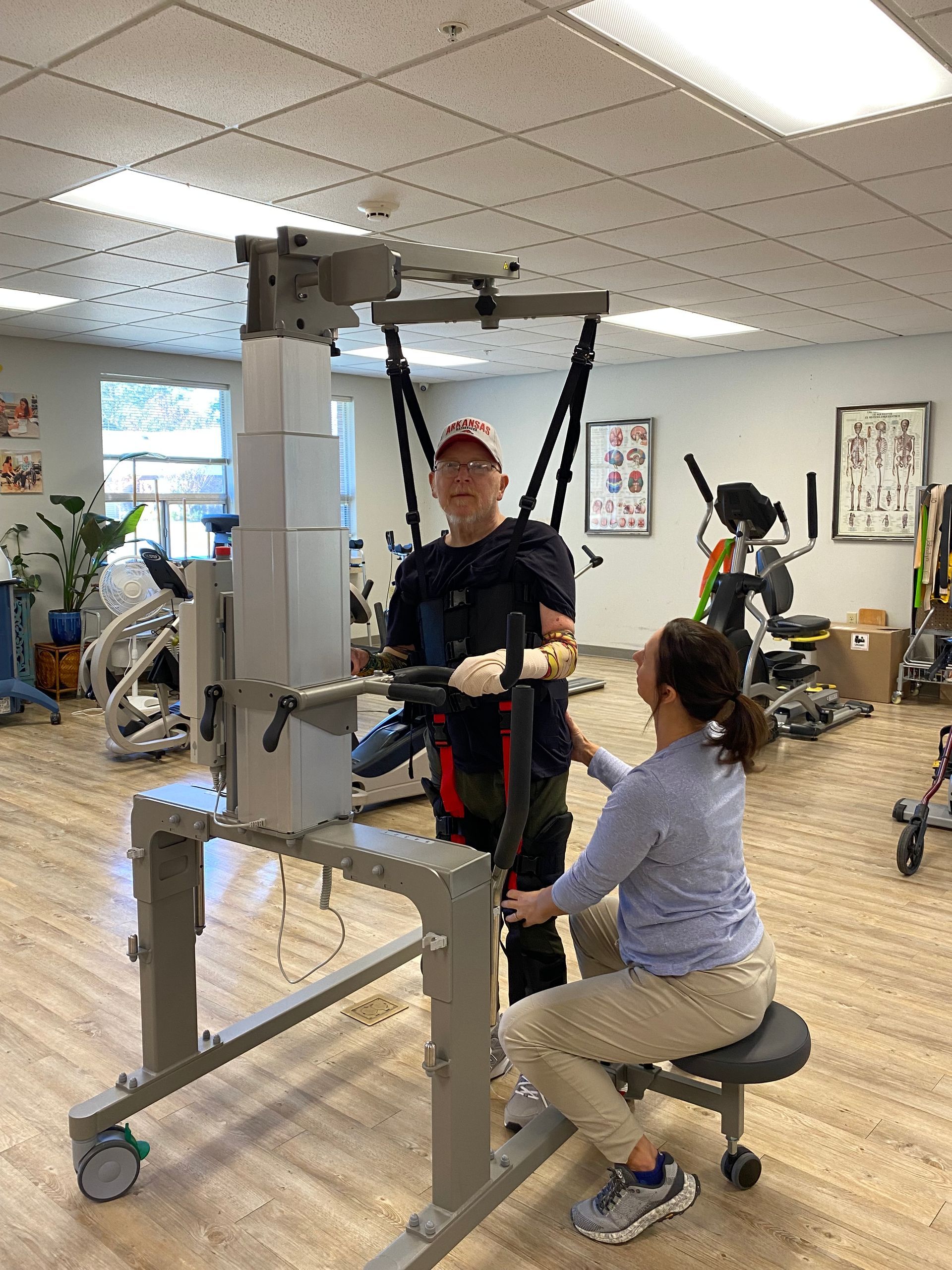 Therapist assisting a patient in a rehab harness beside a seated patient in a physical therapy gym