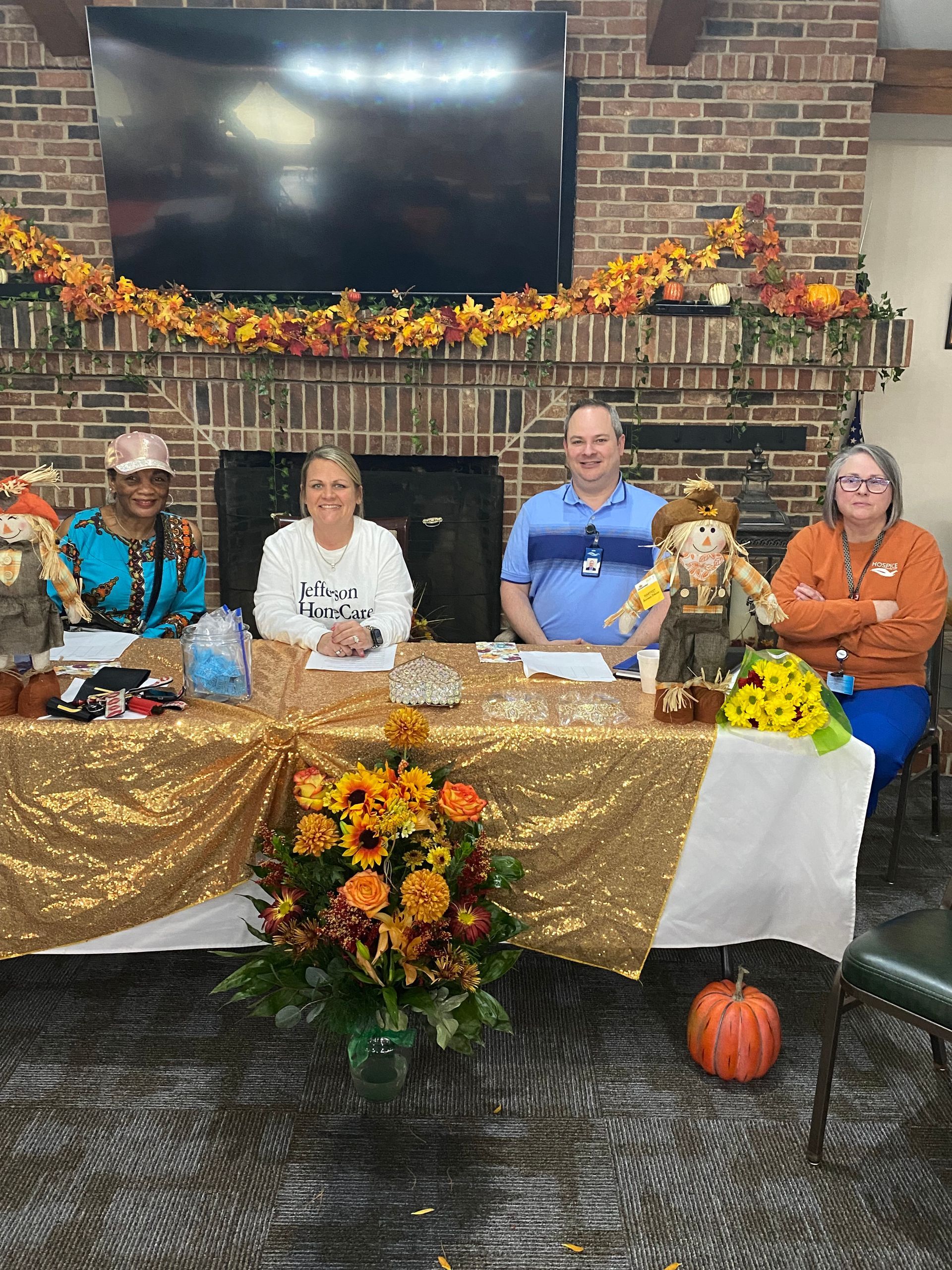 Five people seated at a decorated table with autumn flowers and pumpkins in front of a fireplace and TV.