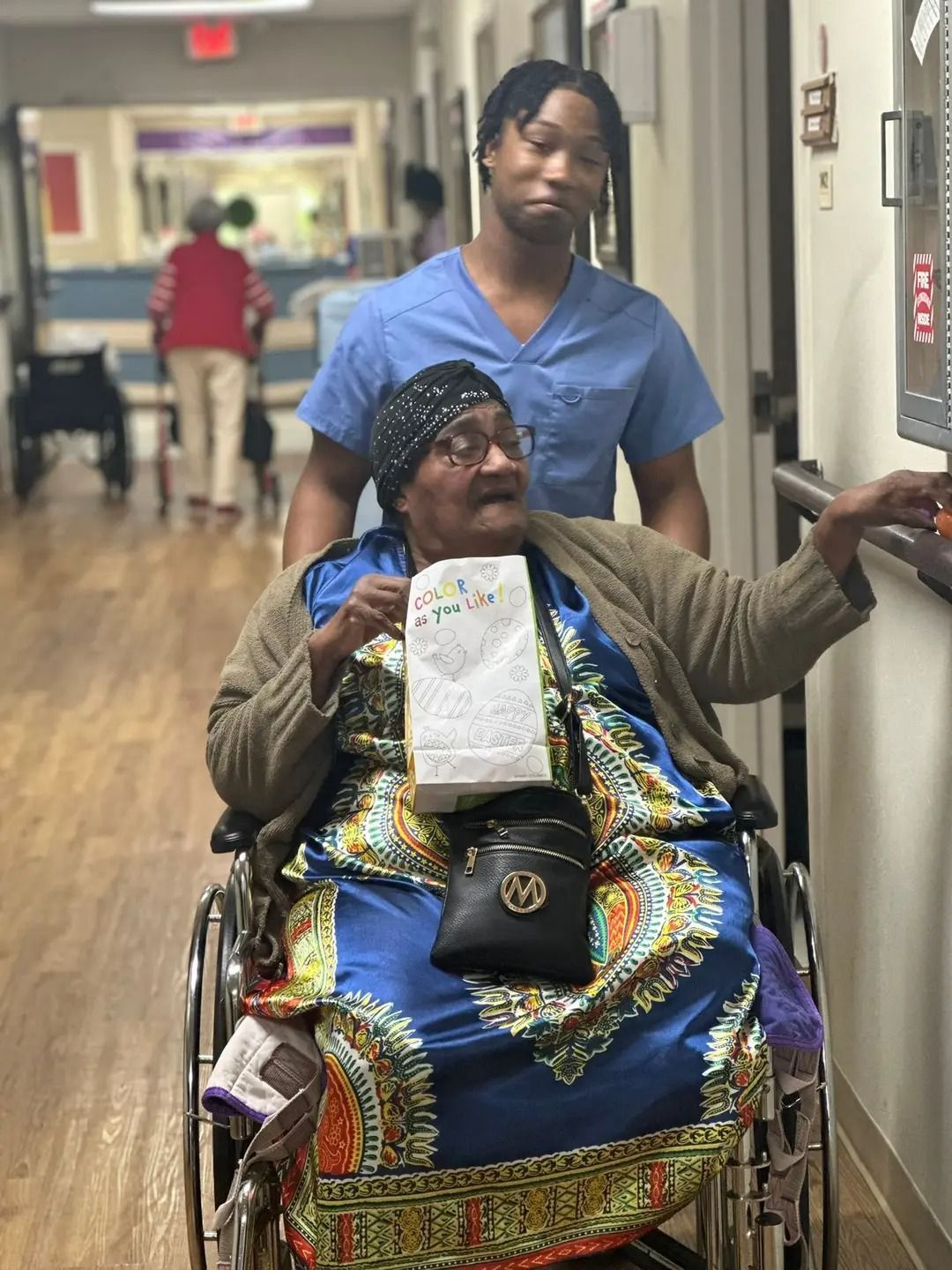 Woman in a wheelchair with a nurse in a hospital hallway, holding a paper and wearing a colorful blanket.