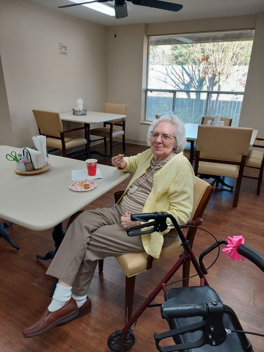 Person seated at a table in a bright dining room, holding a walker, with coffee cups and chairs nearby.
