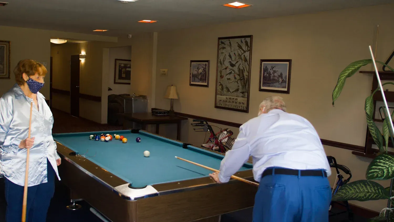 Two people playing pool in a lounge, with framed wall art and a houseplant nearby.