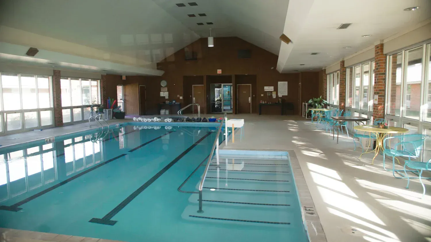 Indoor swimming pool with lanes, windows, and tables along the deck, lit by sunlight
