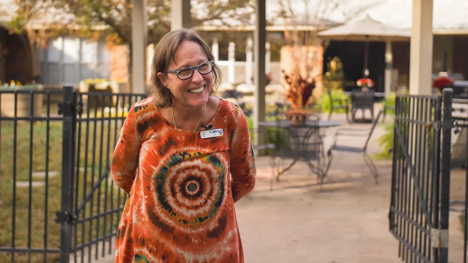 Smiling person in an orange patterned top standing by a gate in an outdoor courtyard
