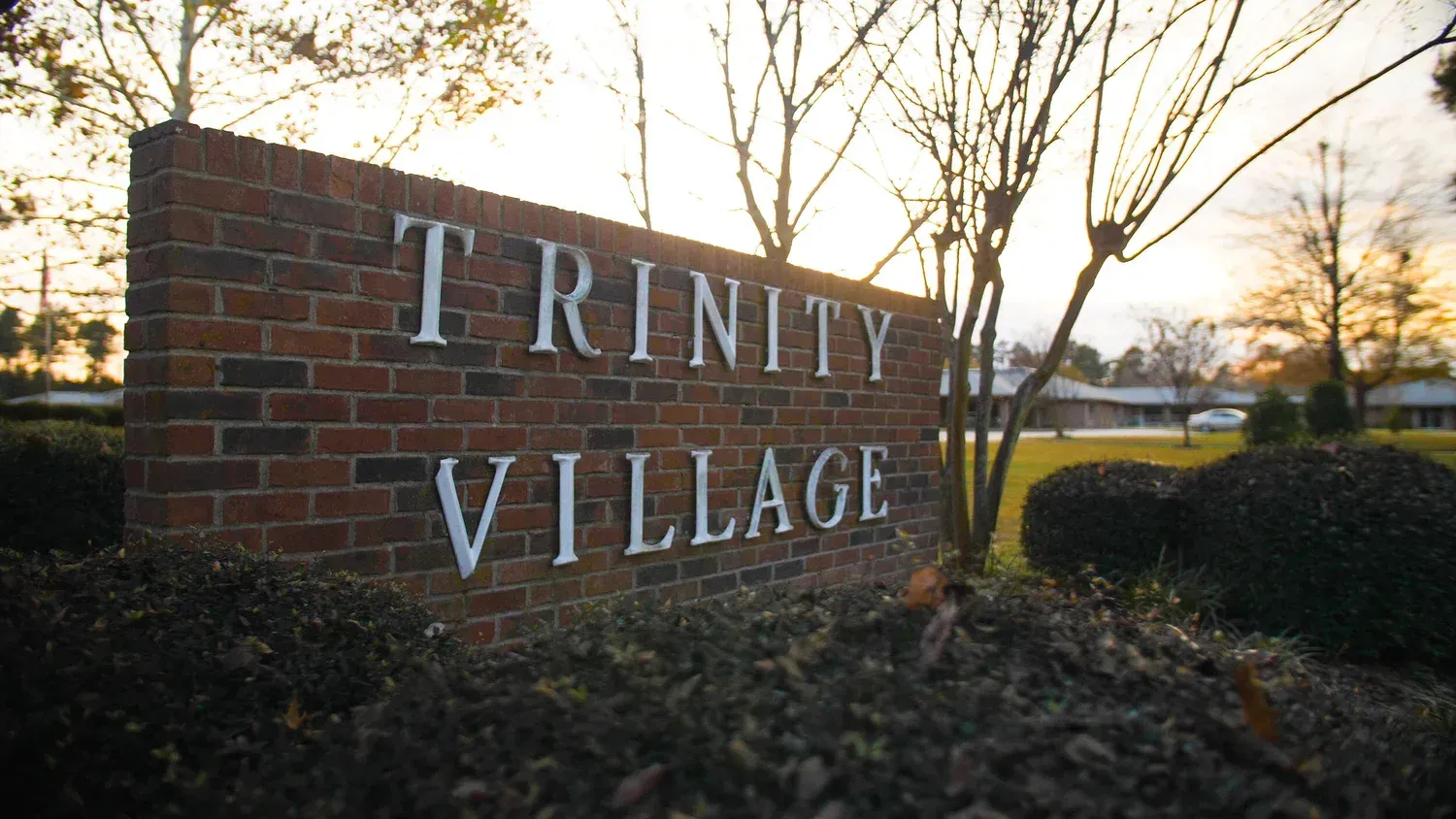 Brick entrance sign reading “Trinity Village” at sunset with trees and landscaping behind it
