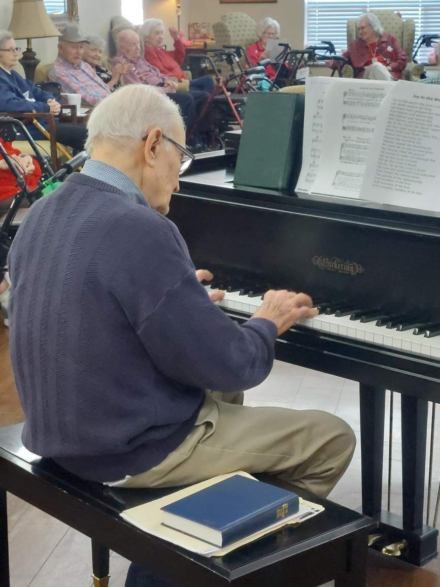 Man playing piano in a community room while singers sit in the background