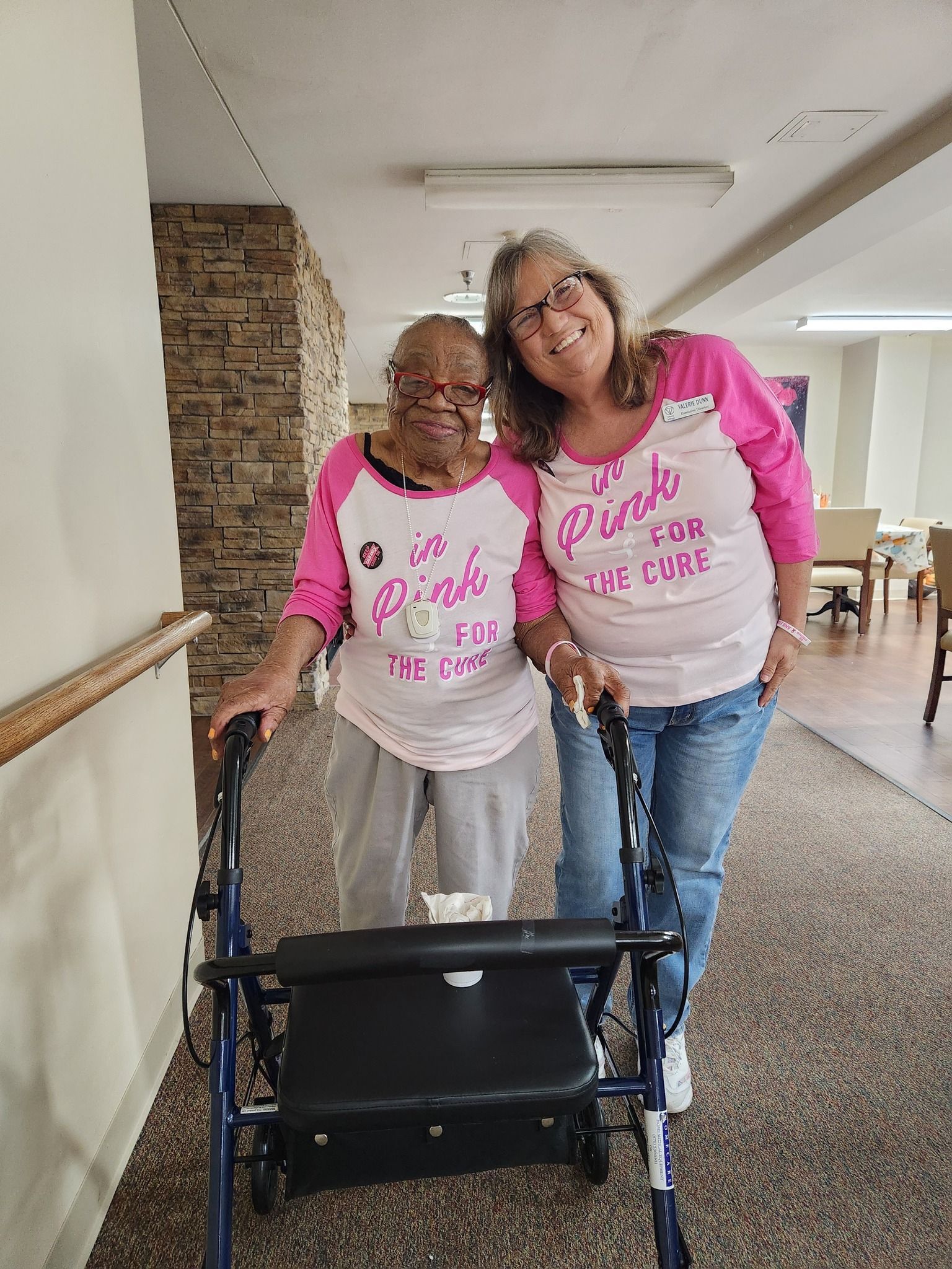 Two women in pink shirts stand behind a black walker in a hallway, smiling and leaning together.