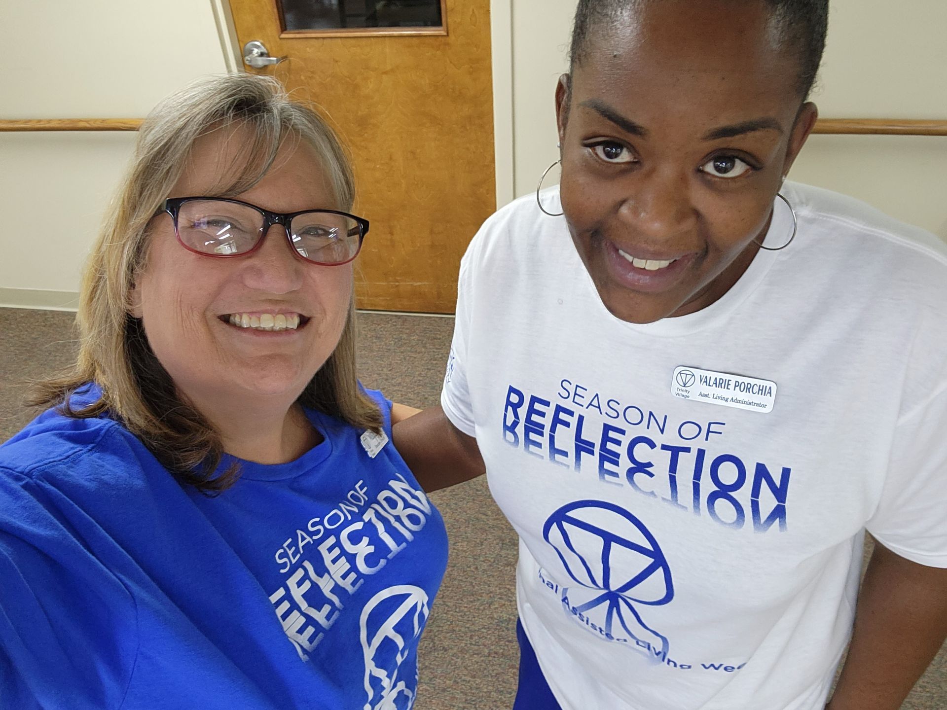 Two smiling people in blue-and-white “Season of Reflection” T-shirts stand indoors.