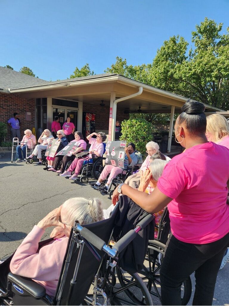 Residents seated outdoors under a covered patio, with a caregiver in pink standing beside them.
