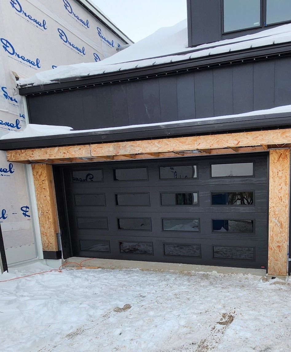 Black garage door on a house under construction, with unfinished wood trim and snow on the ground