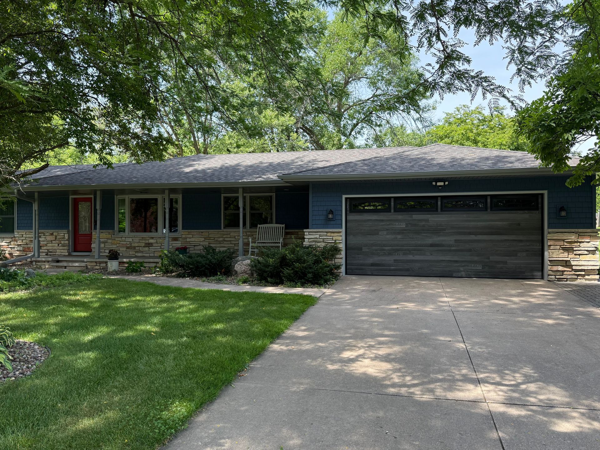 Single-story blue house with a red front door, large garage, and tree-shaded driveway