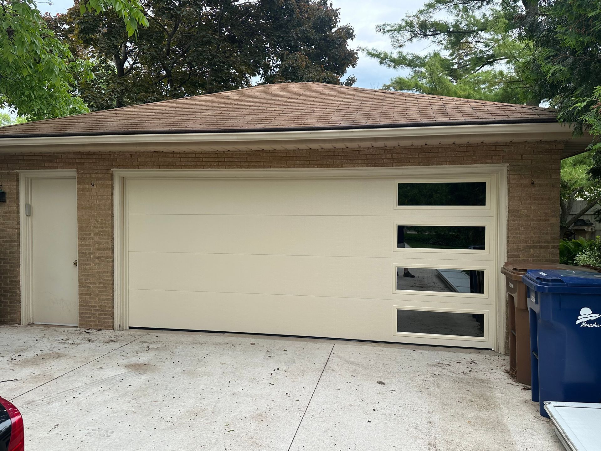 Detached garage with beige door, stone trim, driveway, and blue recycling bins beside it