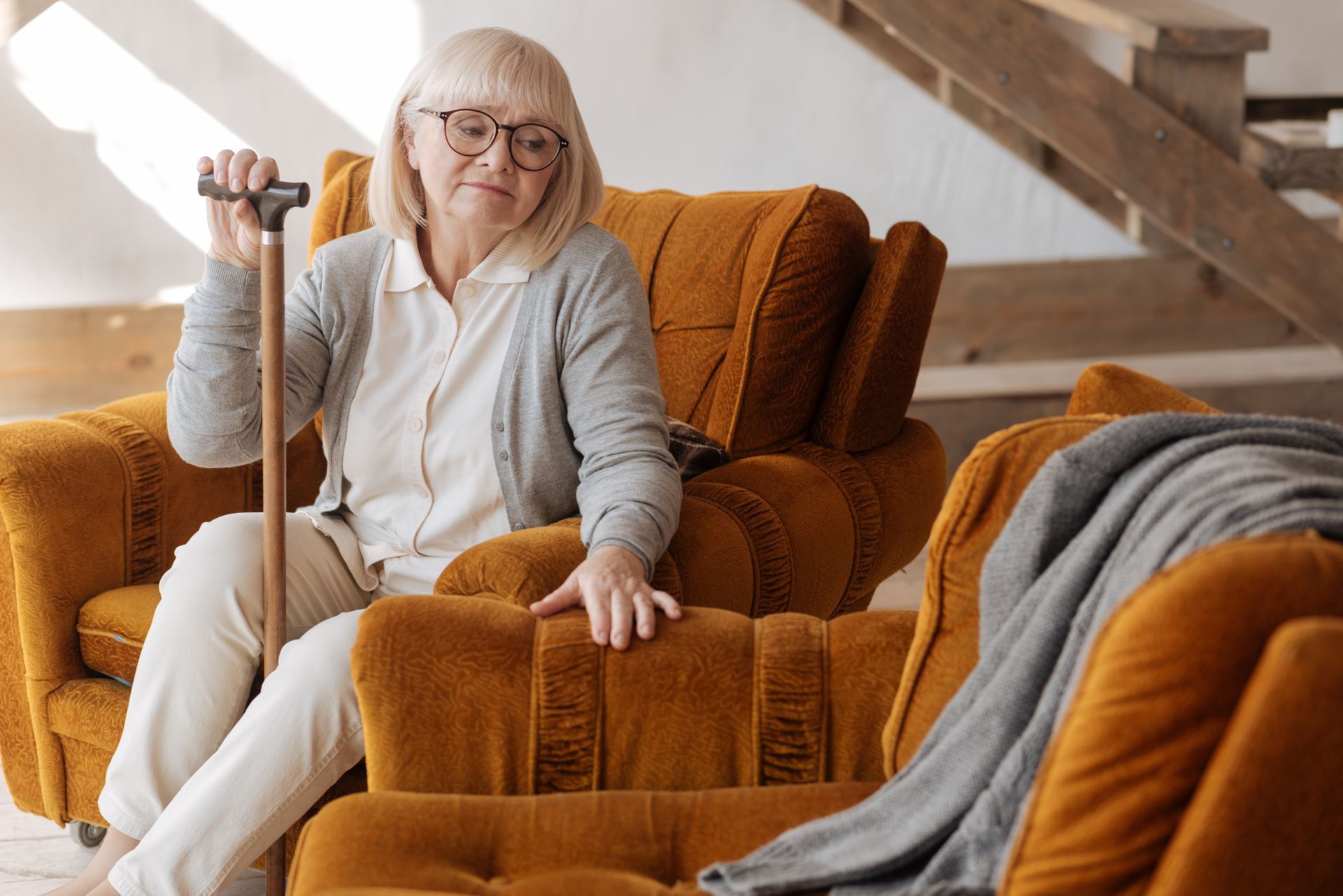 Woman in a gray outfit sitting in an orange armchair, holding a cane in a cozy living room.