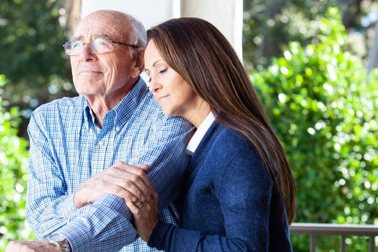 Two people standing together outdoors, holding hands and looking down thoughtfully.