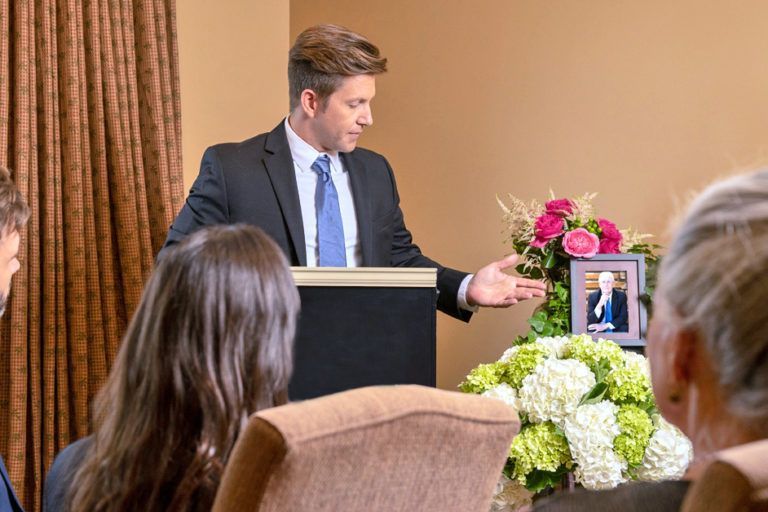 Man speaking at a podium during a memorial service, with flowers and a framed photo nearby