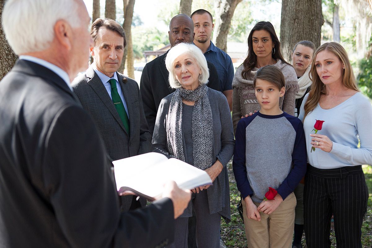 A man speaks to a small outdoor group holding papers under trees, with attentive faces.