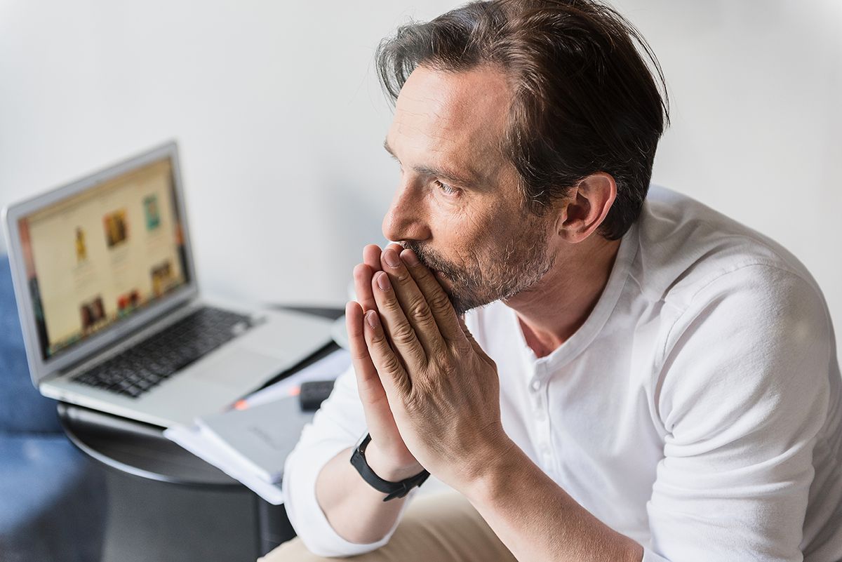 Man in white shirt sitting at laptop, hands clasped near his face, looking concerned