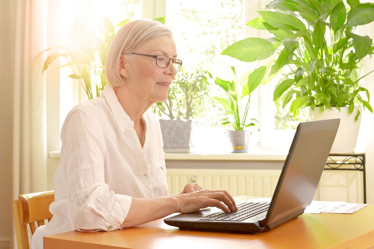 Woman working on a laptop at a sunlit wooden table with green plants in the background