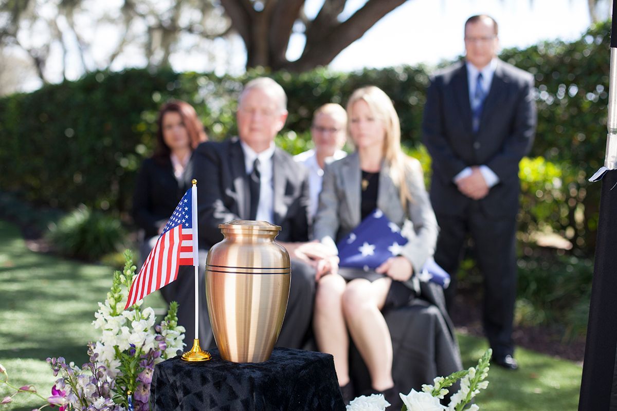 Memorial display with small American flags and urn, mourners seated outdoors in a garden setting